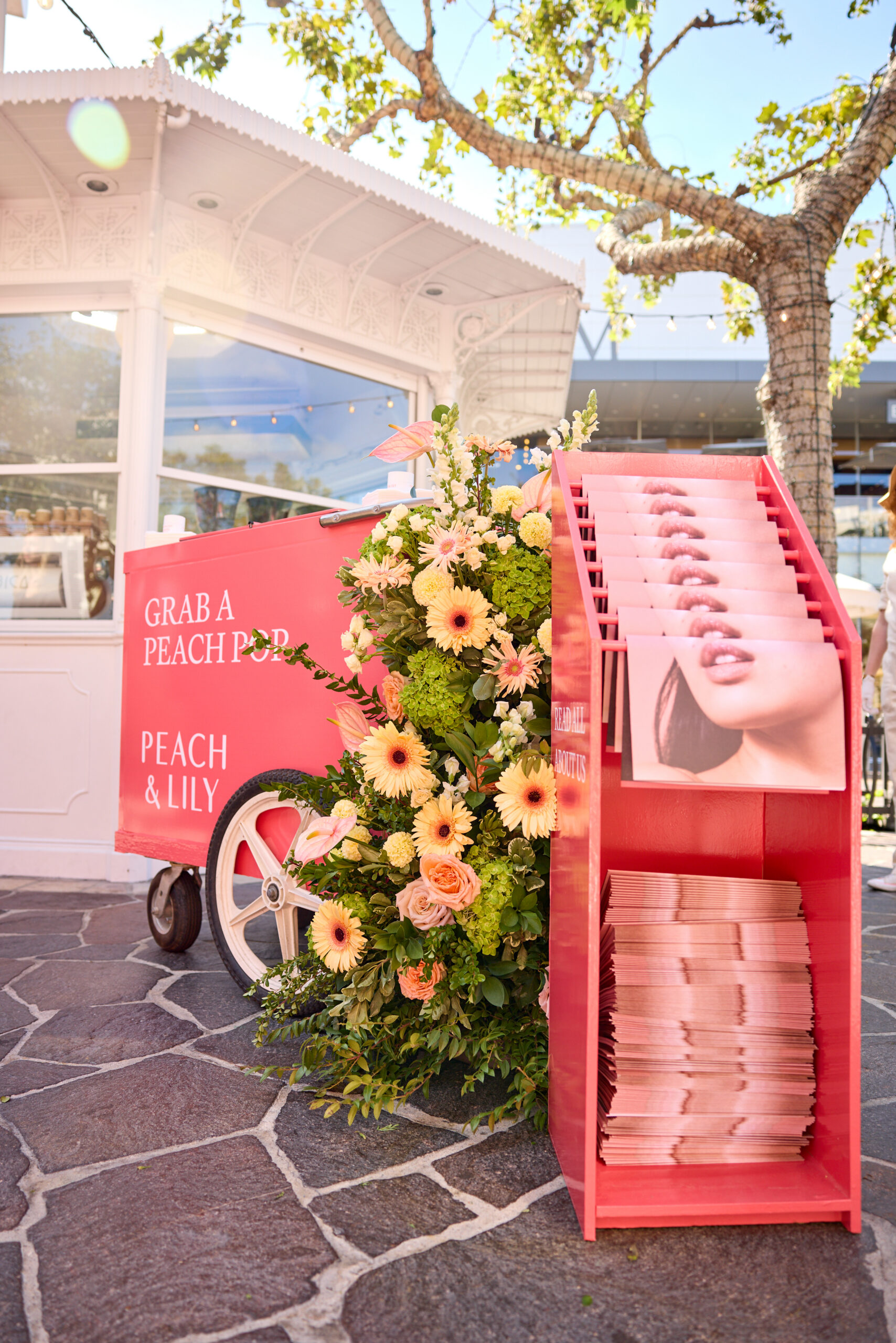 Peach & Lily pop-up bus cart with flowers and pink magazines near a white gazebo outdoors.