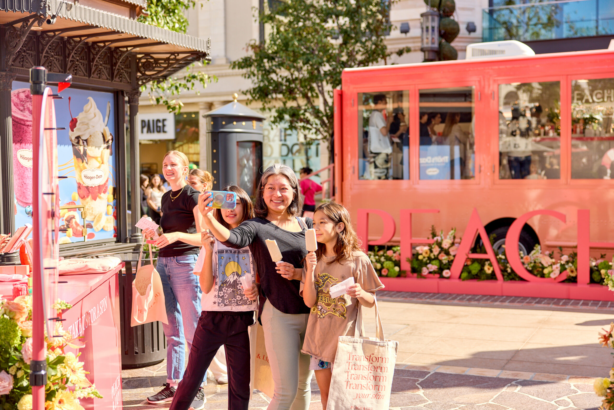Three women take selfies while eating ice cream near a pink Peach & Lily bus at an outdoor event.