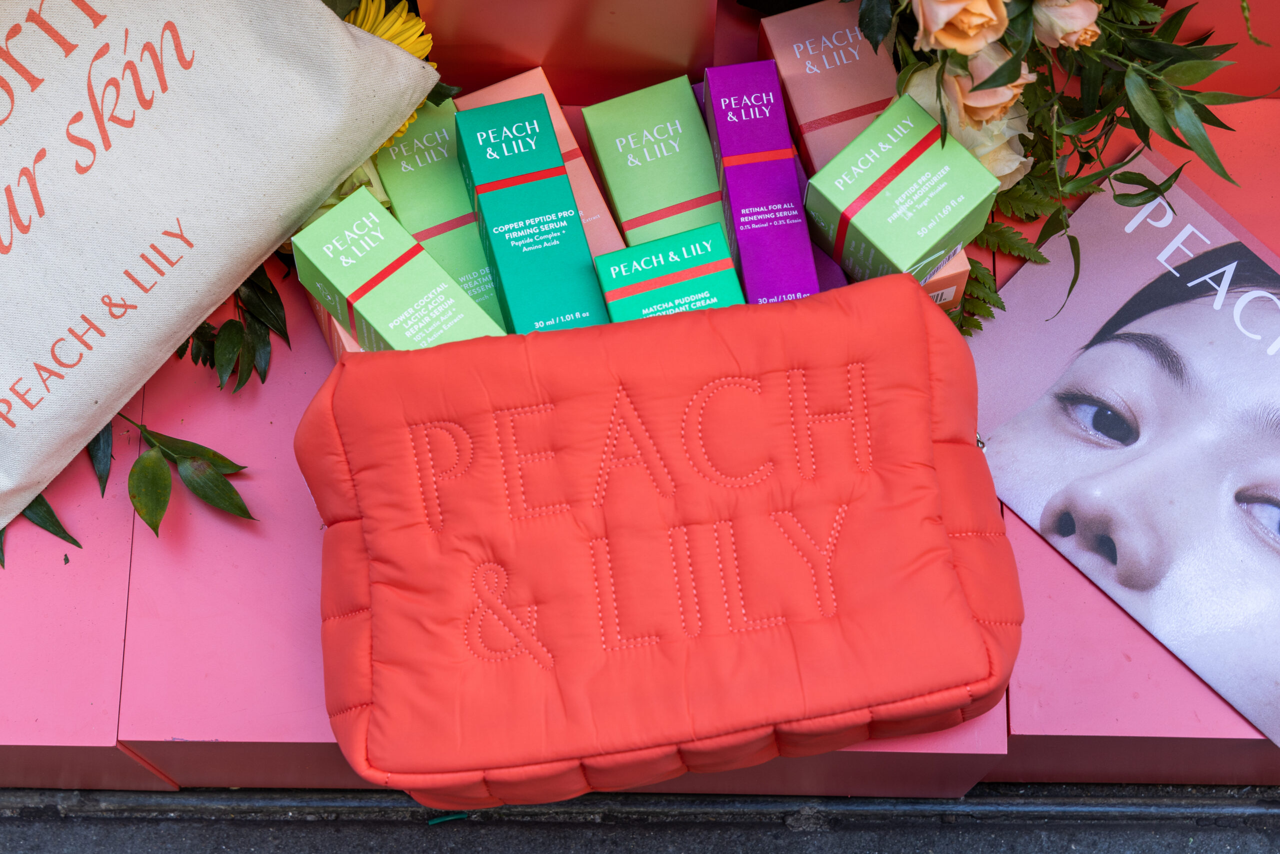 Coral Peach & Lily pouch with skincare boxes, flowers, and part of a woman's face on a magazine.