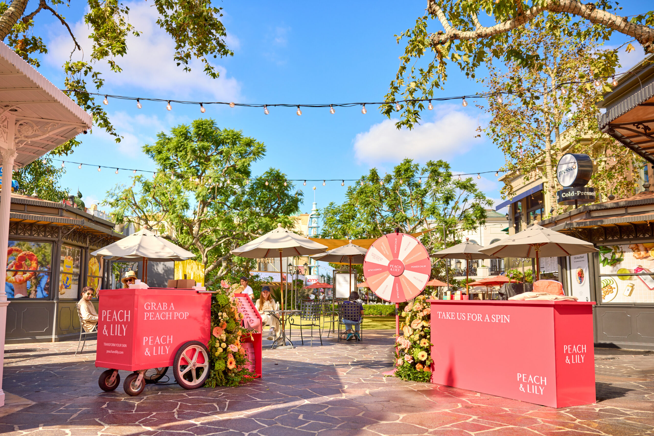 Outdoor plaza with pink Peach & Lily booths, prize wheel, pop-up bus, and shoppers browsing.