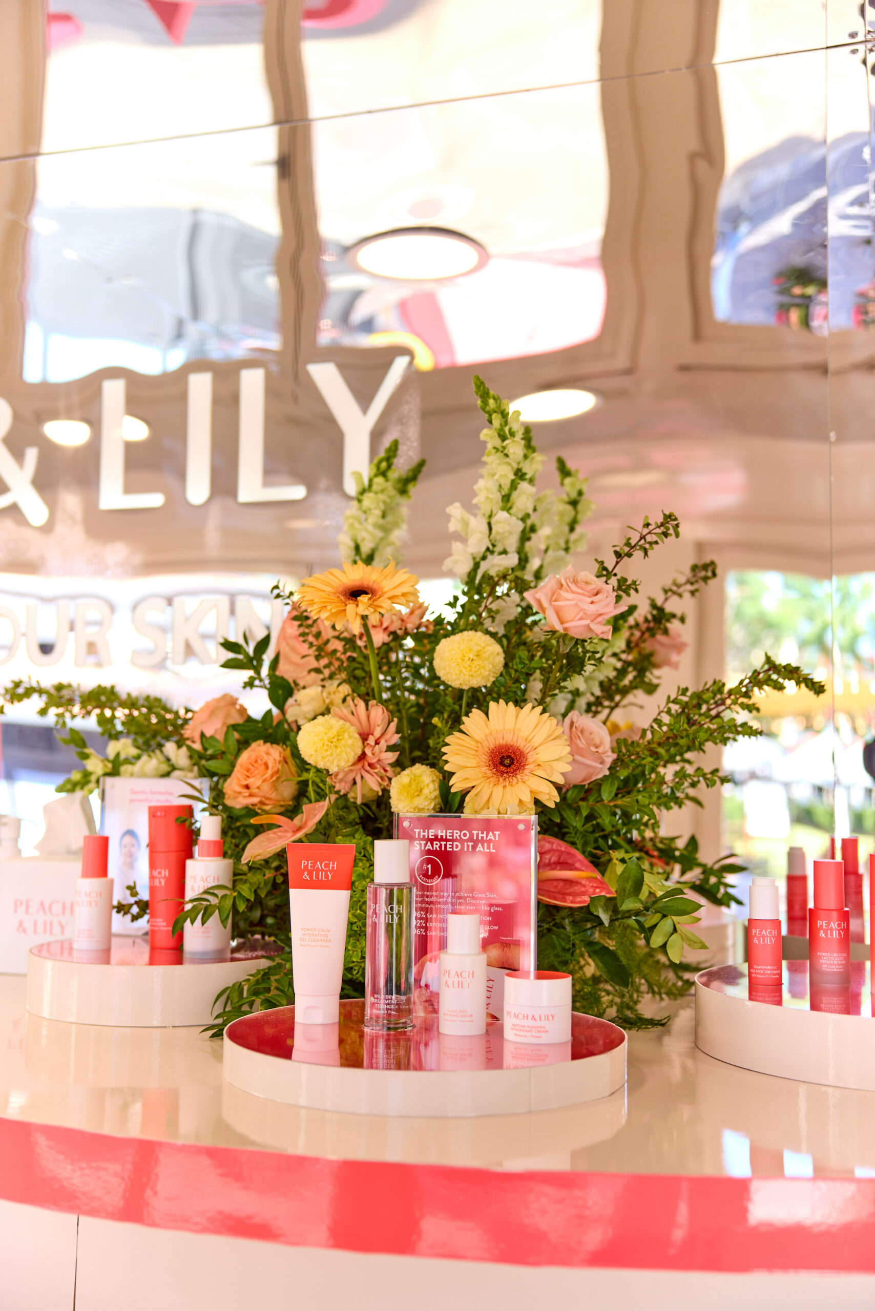 Peach & Lily skincare on trays with colorful flowers, set before a mirrored branded backdrop.