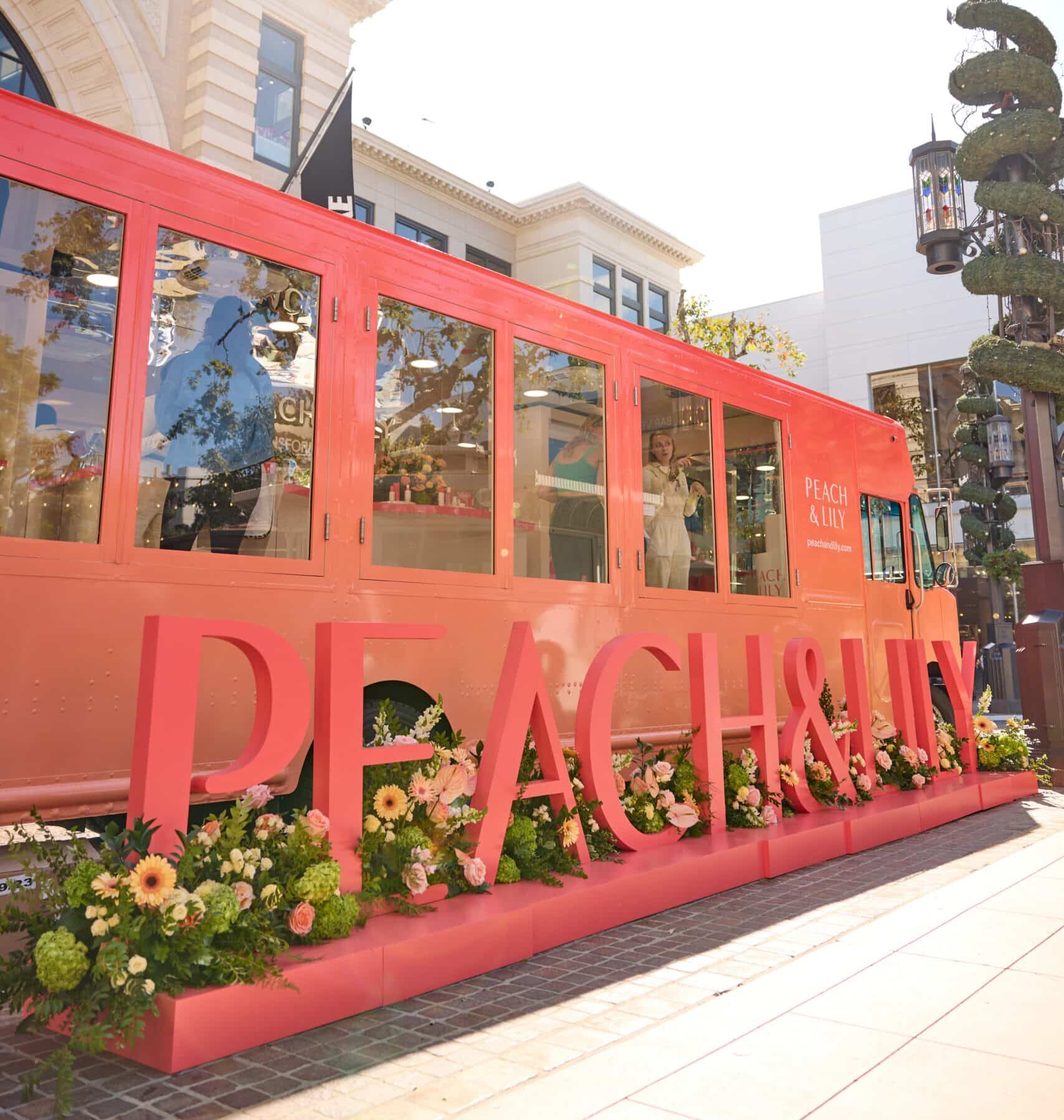 Pink Peach & Lily pop-up bus with large lettering and flowers parked outside near buildings.