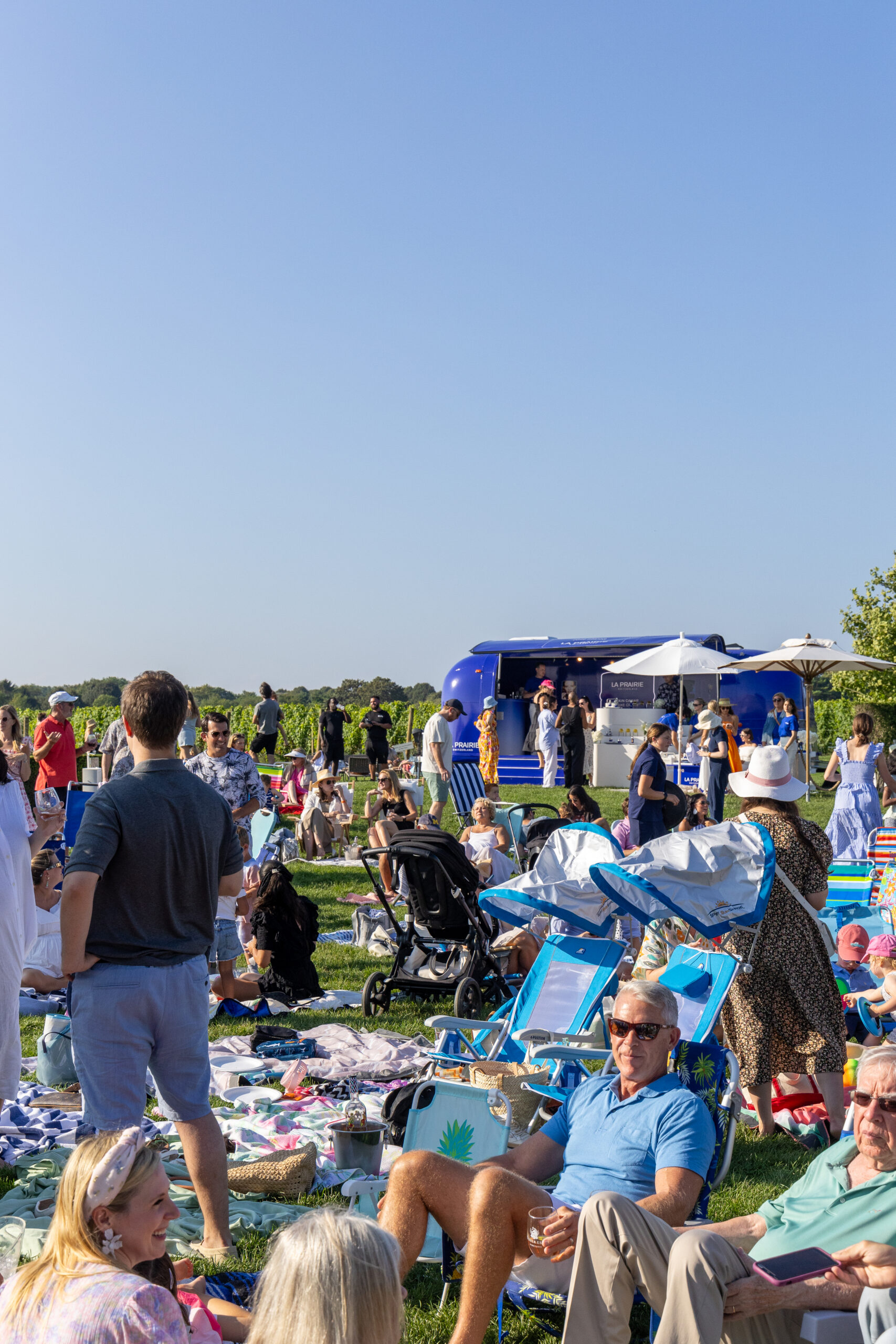 People sit on grass at an outdoor event, relaxing near a blue food truck and La Prairie pop up.