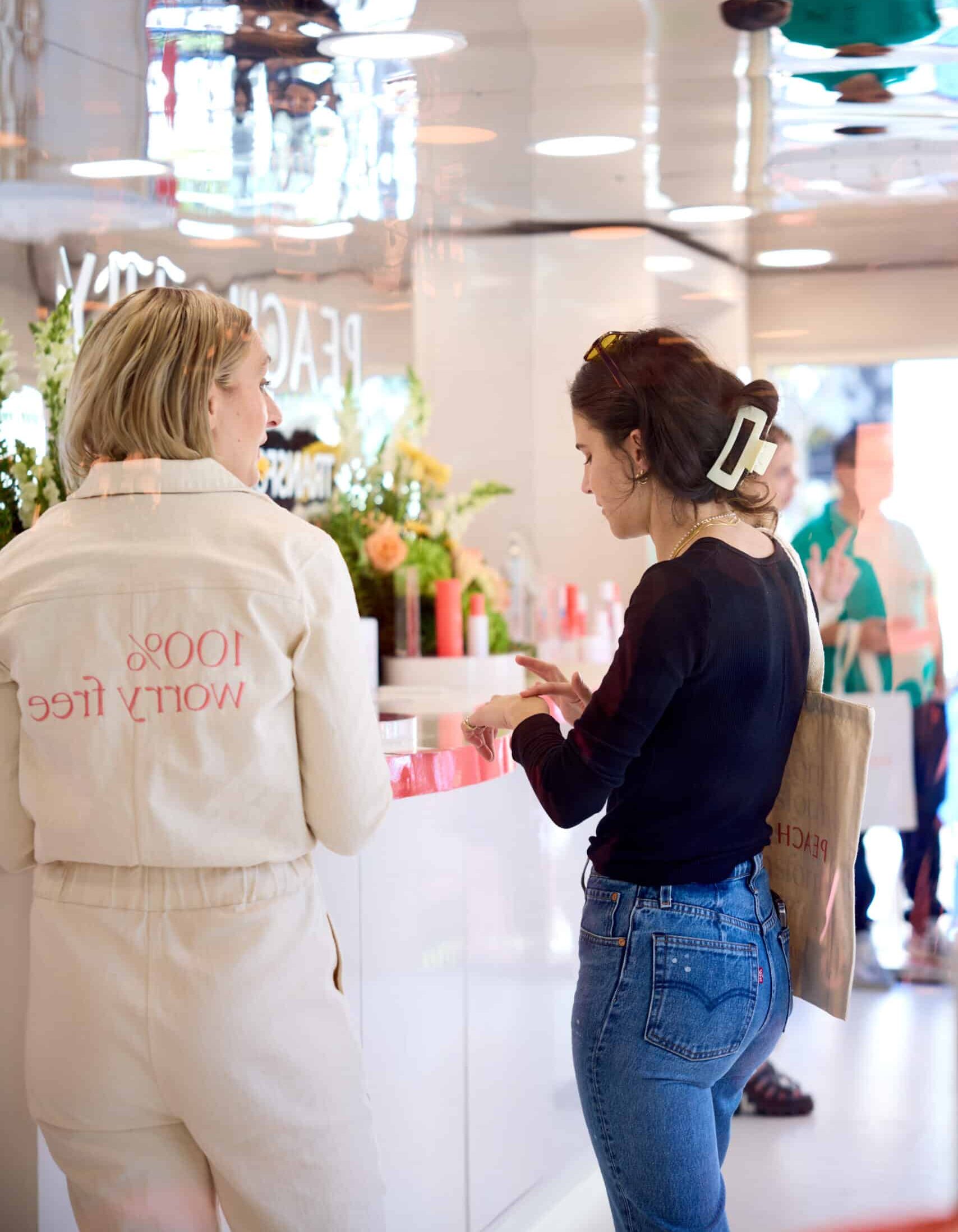 Two women talk inside a Peach & Lily pop-up bus at a white counter with flowers.