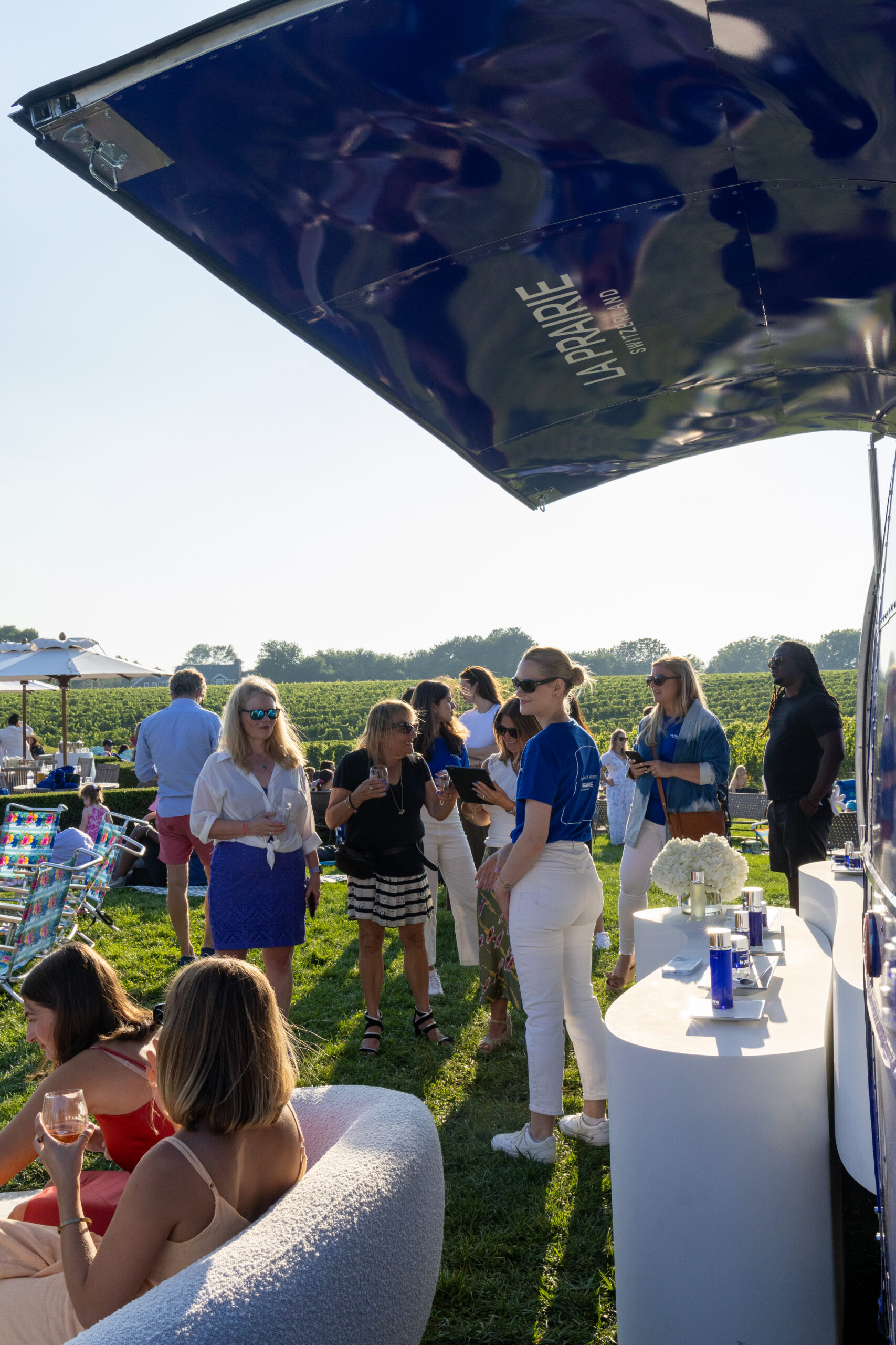 People socialize near a blue and white la prairie pop up bar with vineyards in the background.