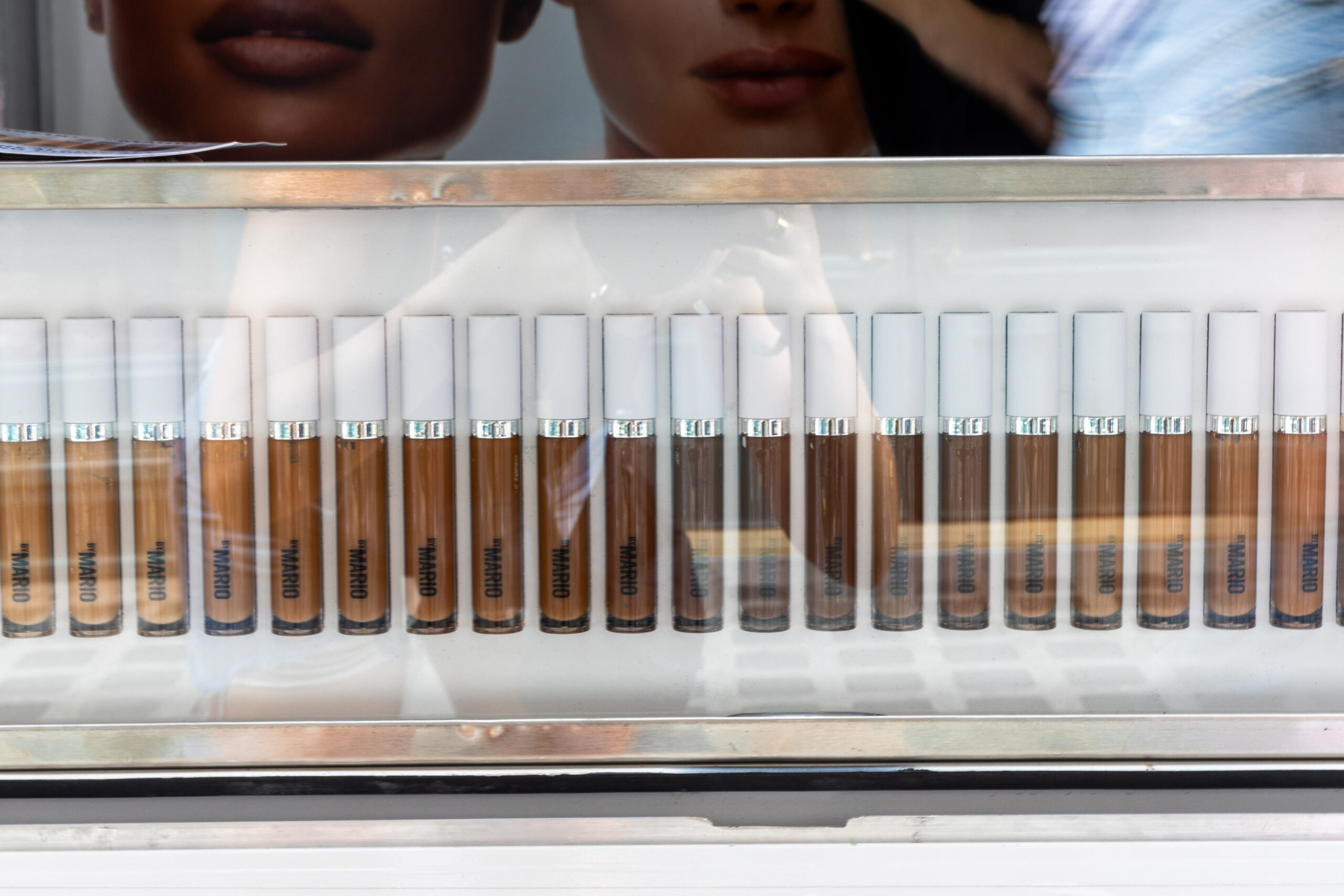 Makeup by Mario foundation tubes in various shades behind glass with people’s reflections visible.