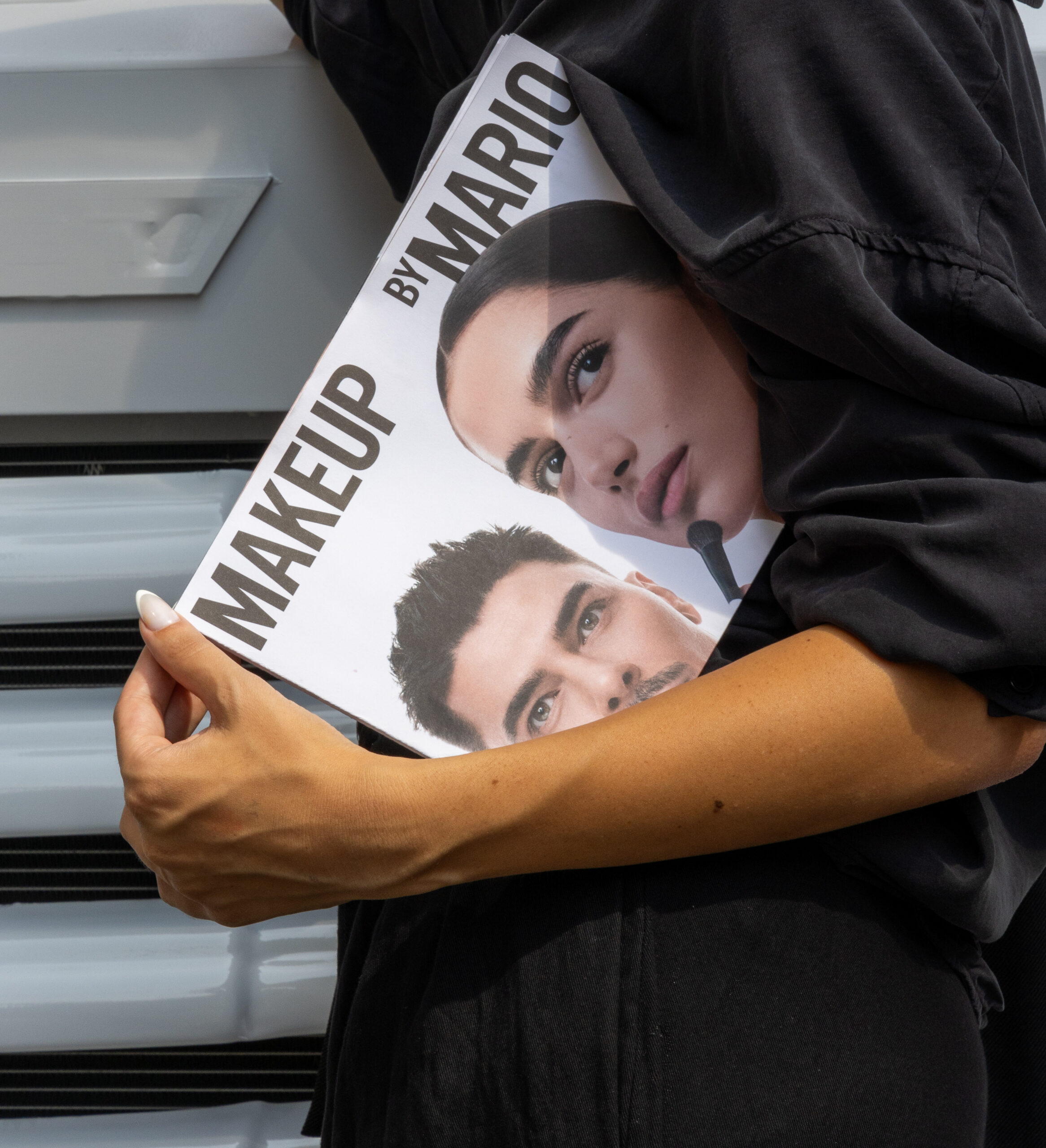 Person in black holds a Makeup by Mario booklet showing close-ups of a woman and a man.