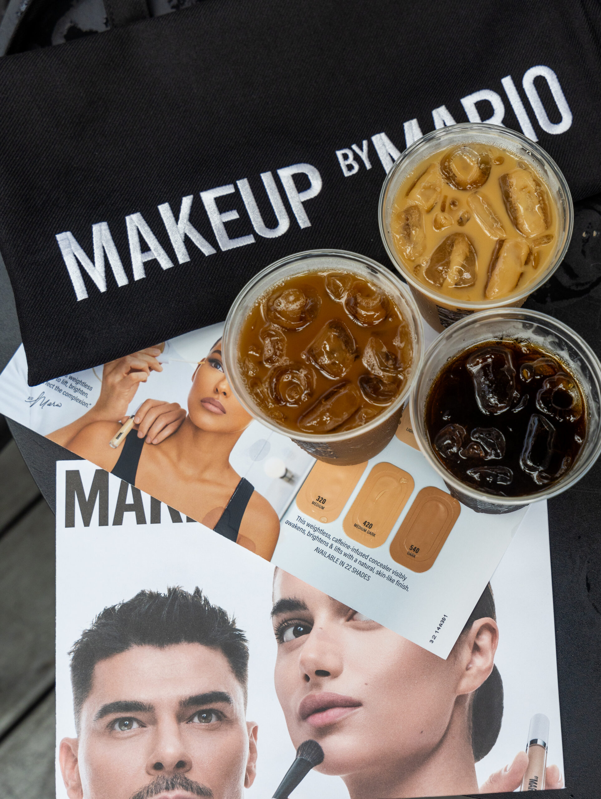 Three iced coffees, a MAKEUP BY MARIO pouch, and makeup ads with models and swatches on a table.