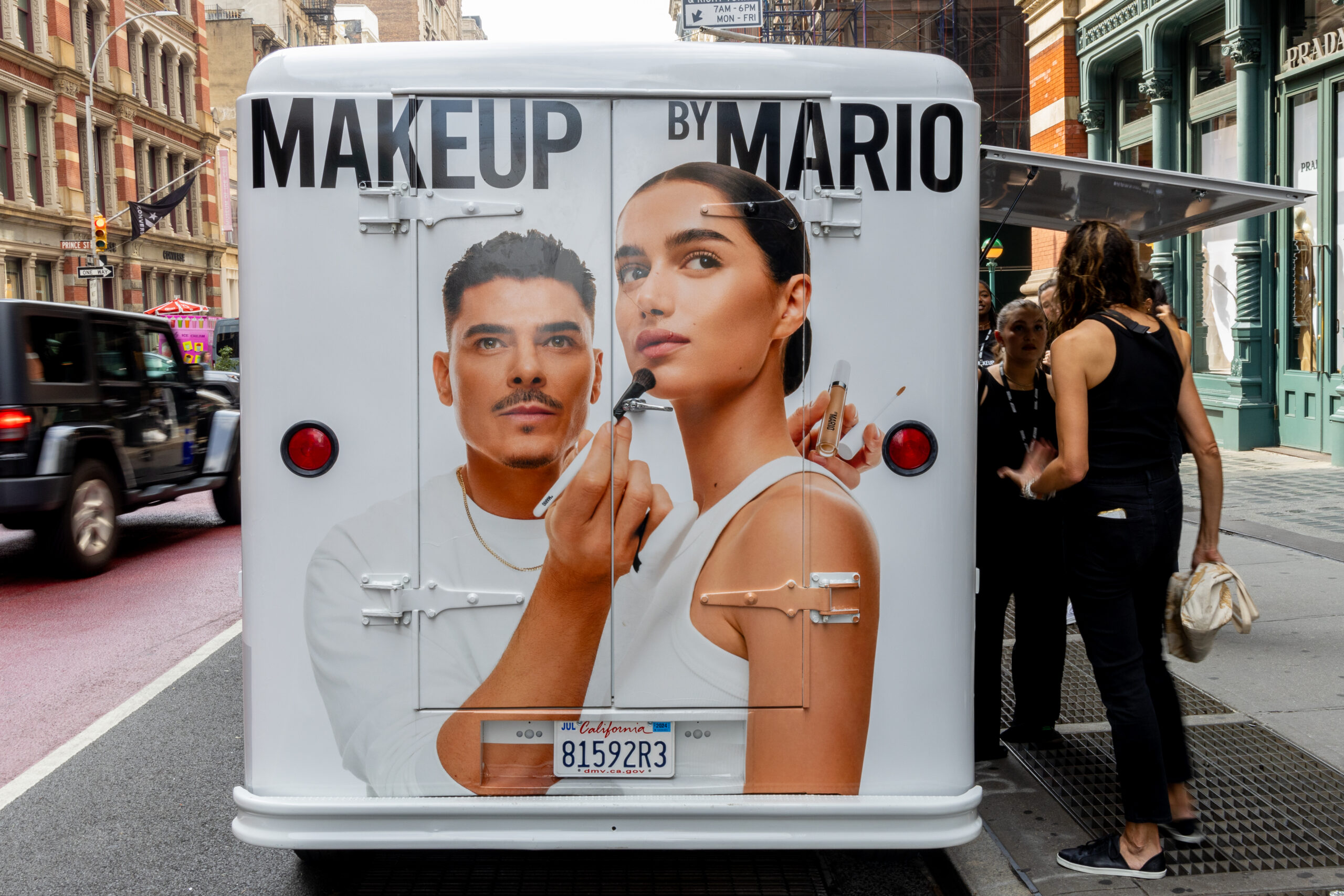 A Makeup by Mario ad truck with two people posing is parked on a city street as others look on.