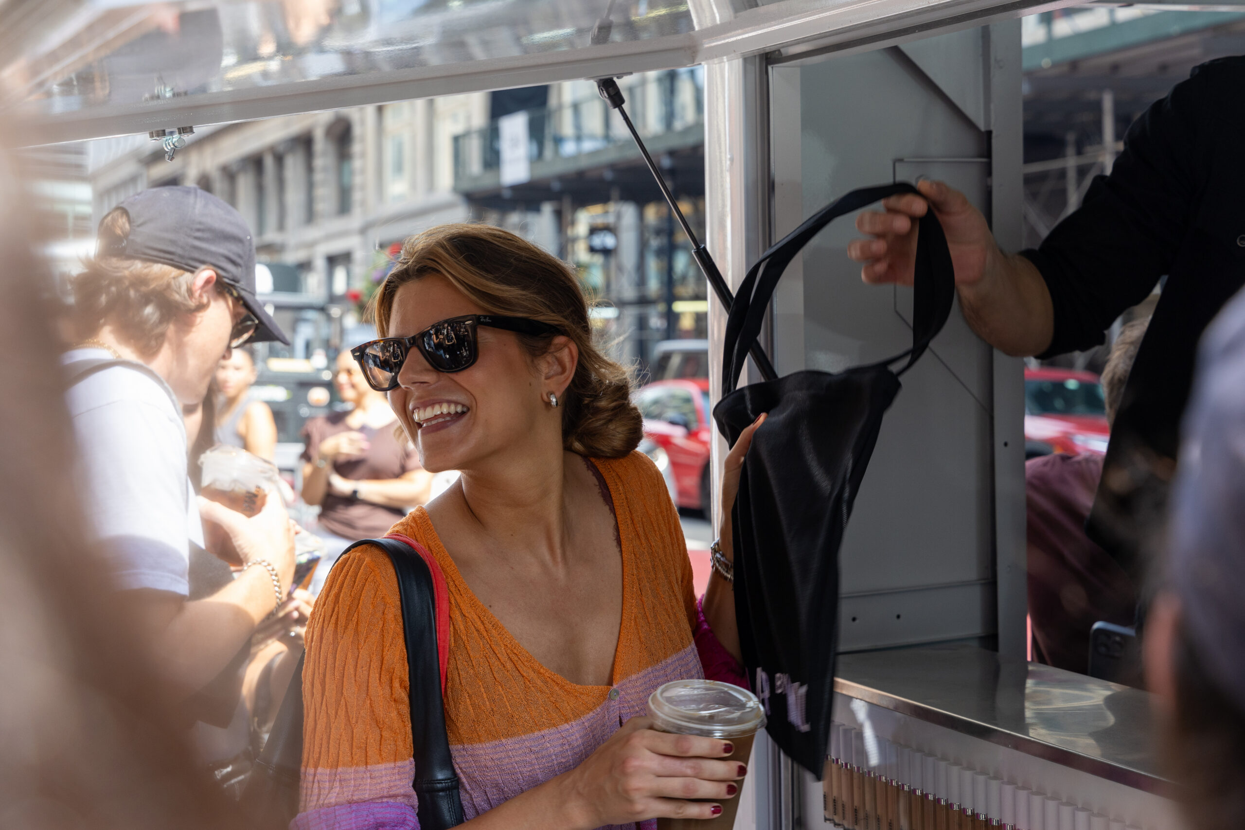 Woman in sunglasses holds a drink and gets a black tote bag from a vendor at an outdoor stand.