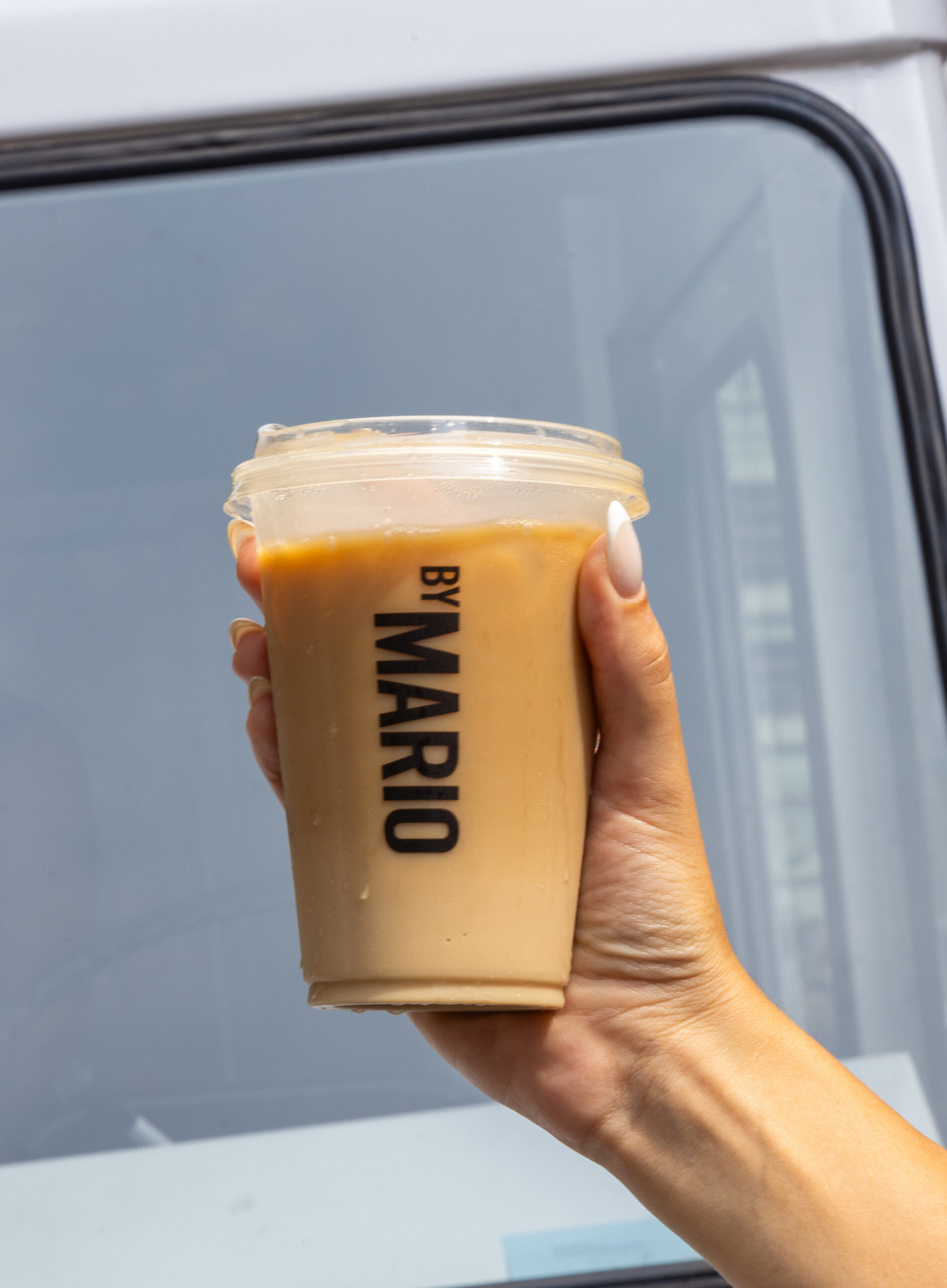 Hand with manicured nails holds an iced coffee cup labeled BY MARIO by a window.
