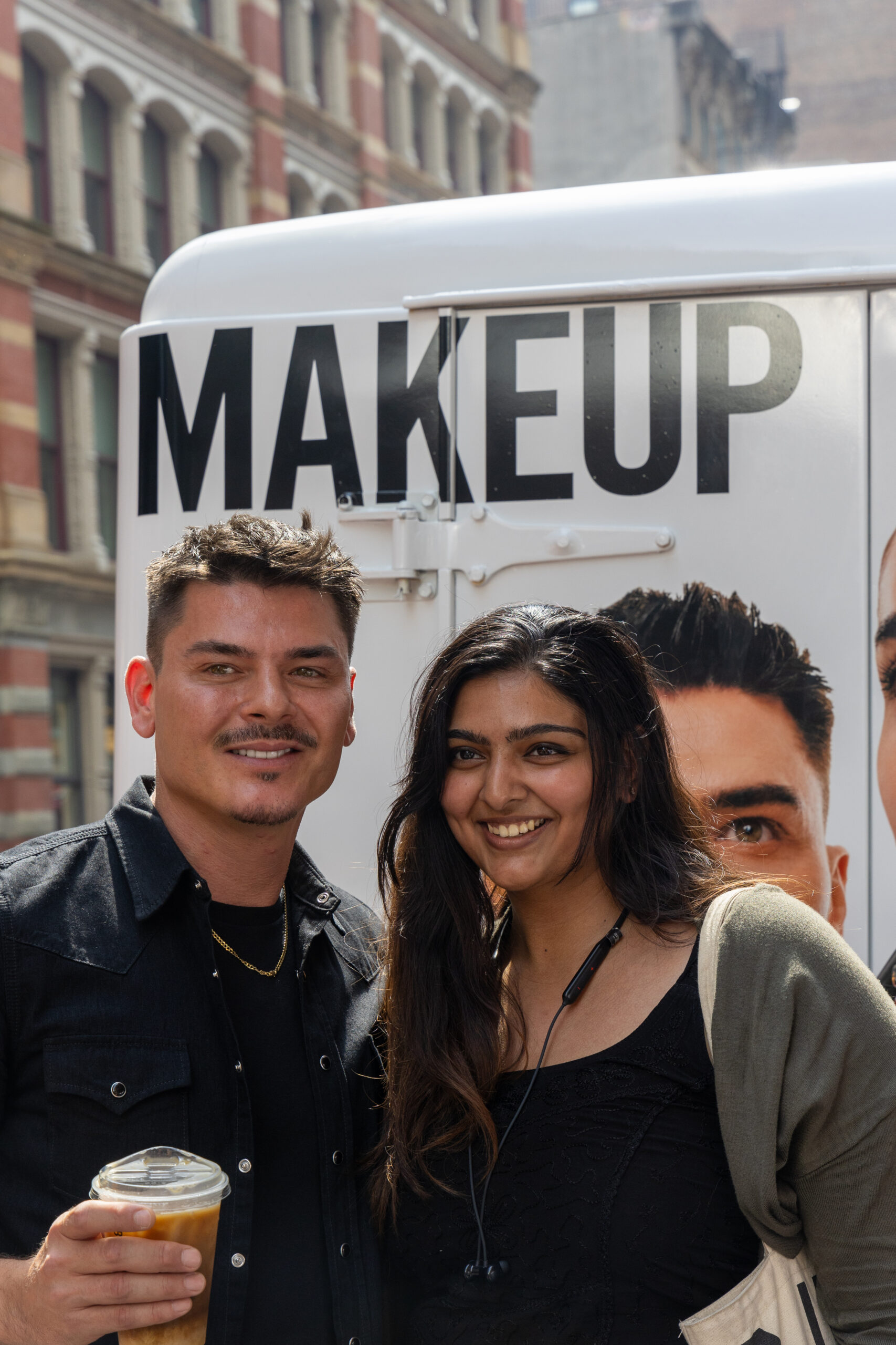 Two people pose and smile in front of a vehicle with a MAKEUP sign; one holds an iced drink.
