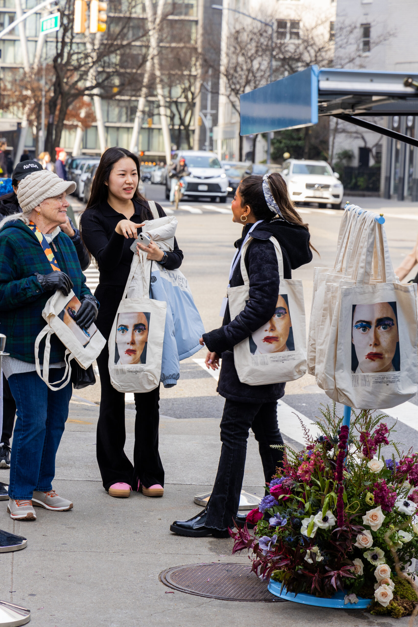 Three women browse tote bags by a peach & lily pop-up bus; flowers sit next to the display.