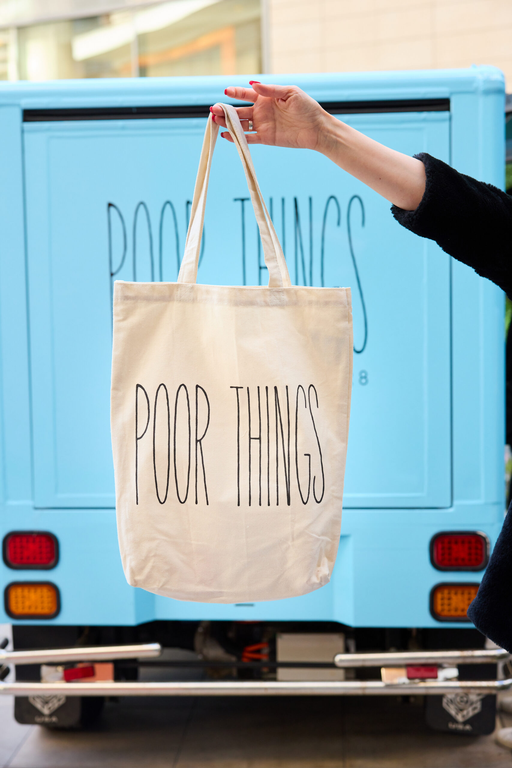 Hand holding beige tote bag with POOR THINGS text, in front of a light blue pop-up bus.