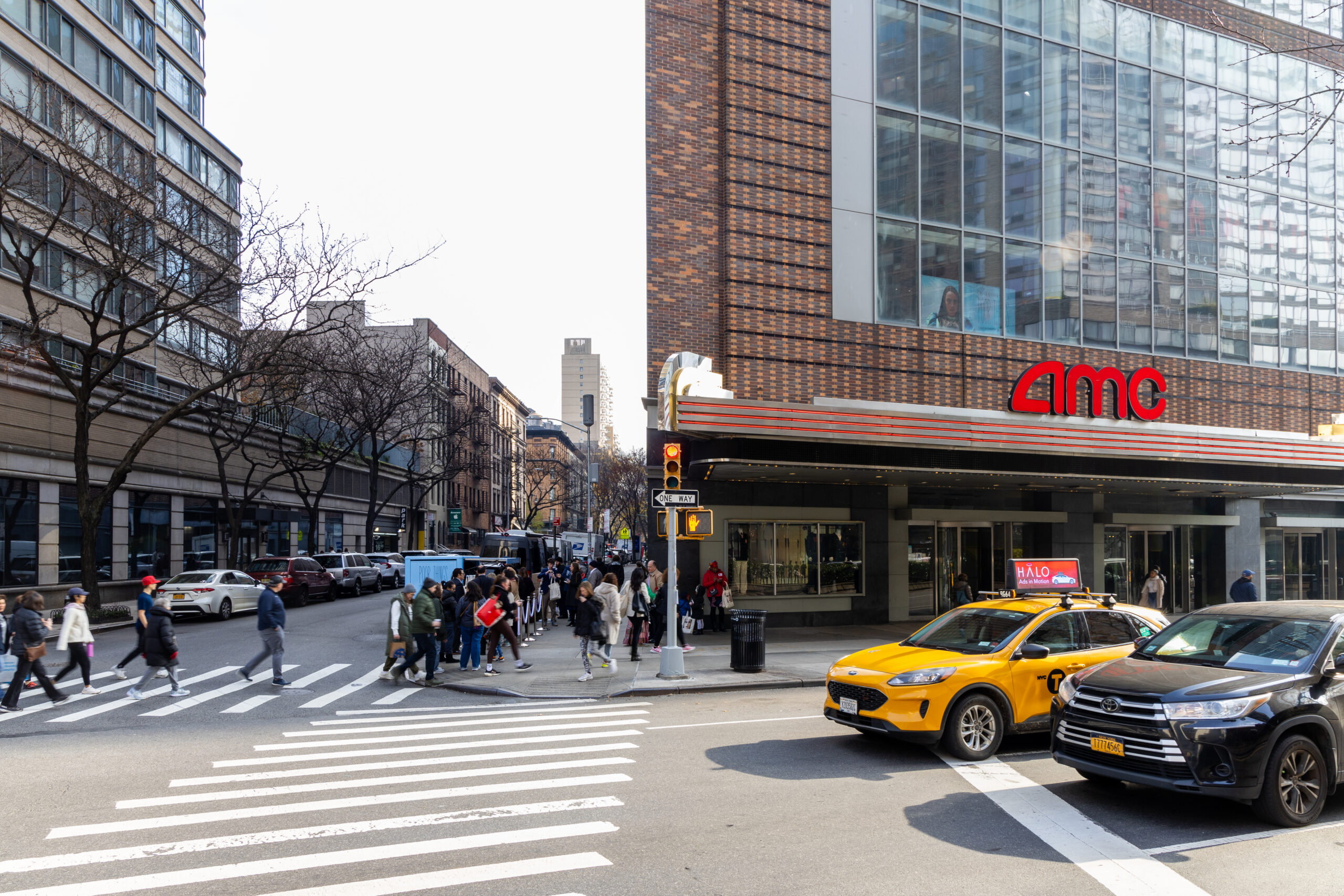 People cross a city street by an AMC theater, with a peach & lily bus and yellow taxis nearby.
