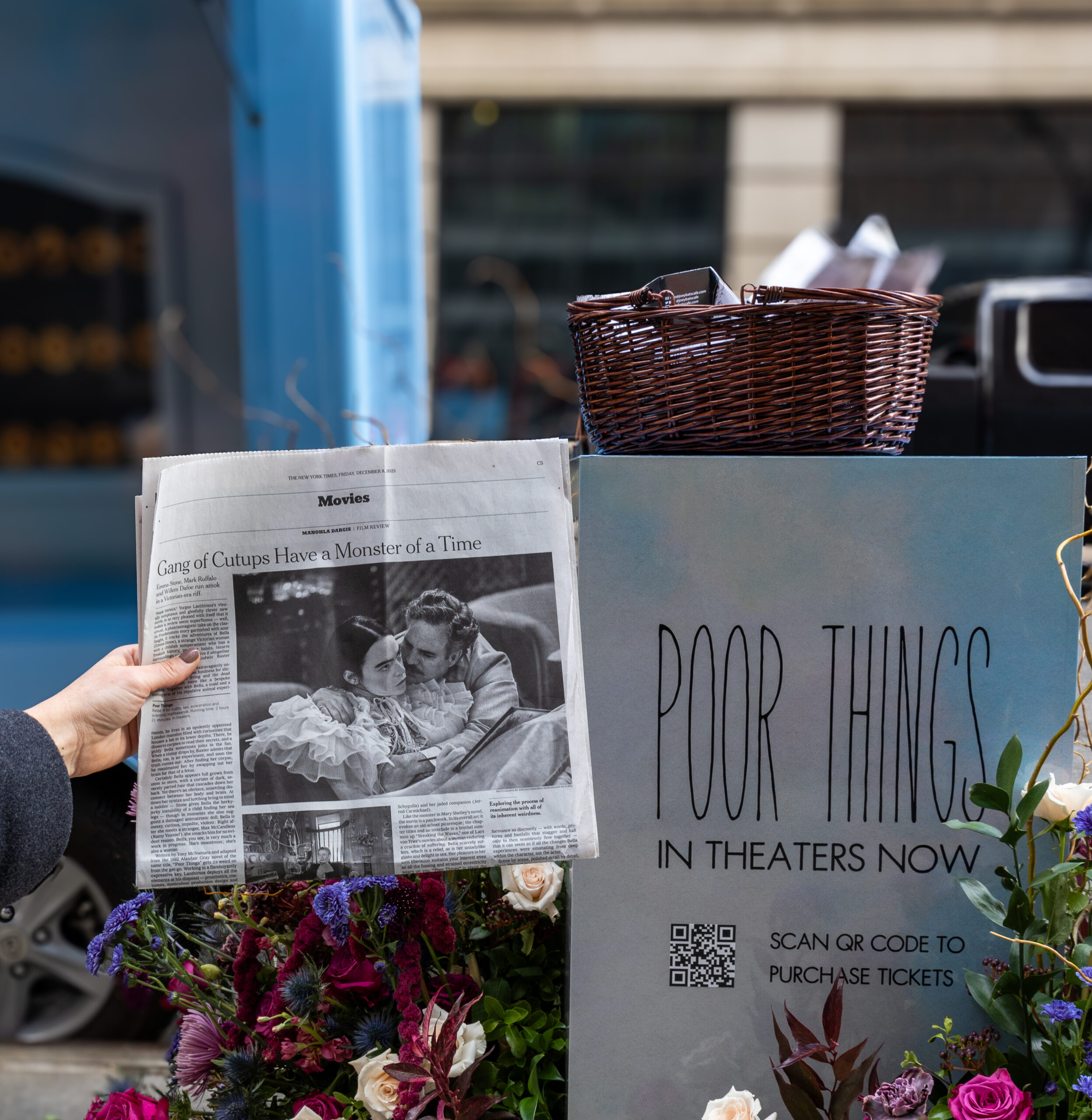 Person holding newspaper with a "Poor Things" photo beside a release sign, flowers, and pop-up bus.