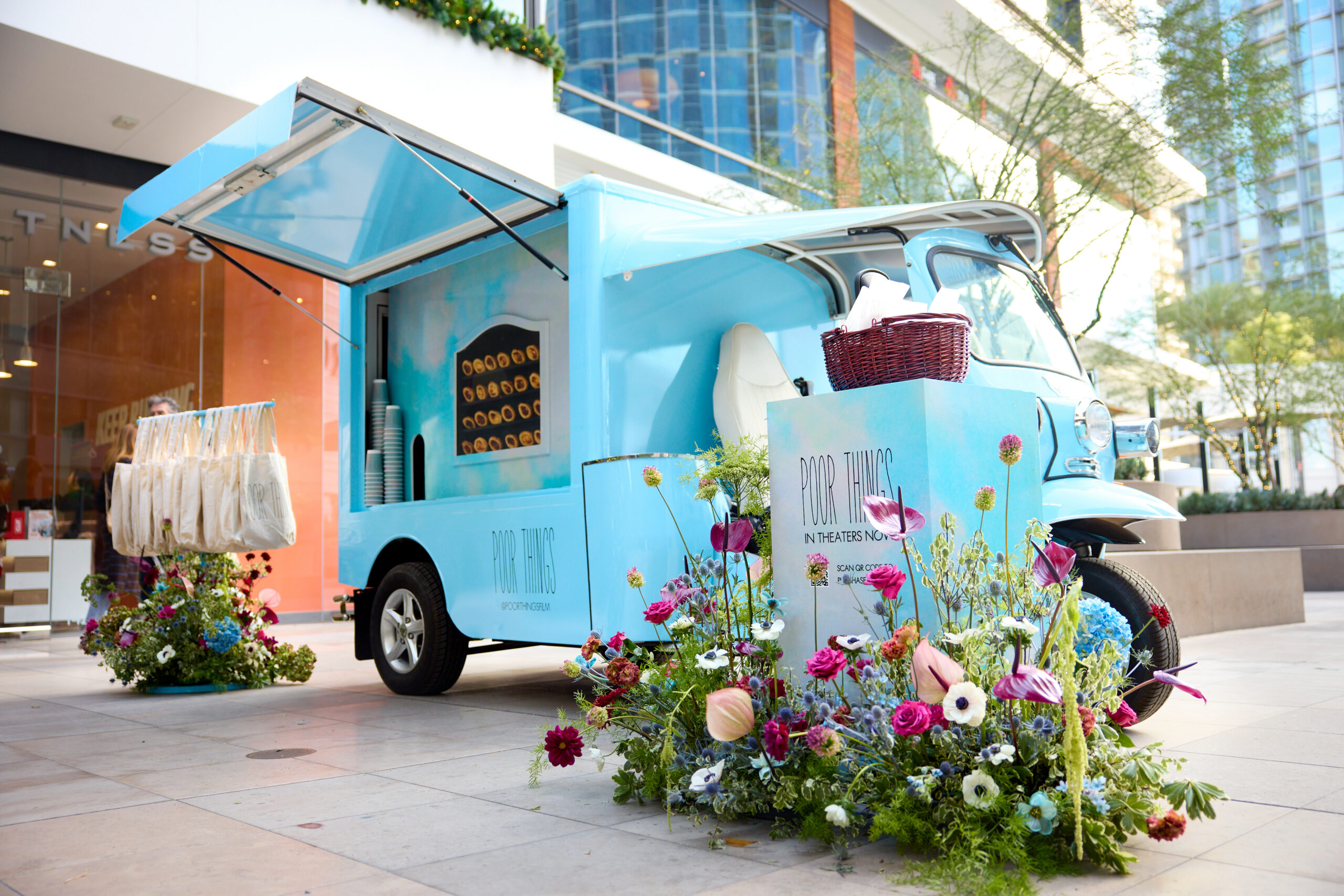 Light blue Peach & Lily pop-up bus with flowers and tote bags parked by modern buildings.