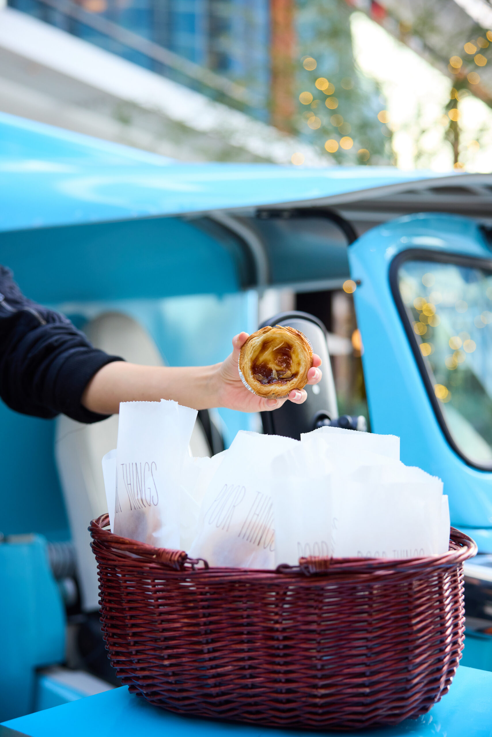 Person holds Portuguese custard tart by a blue Peach & Lily bus and a basket of paper bags.
