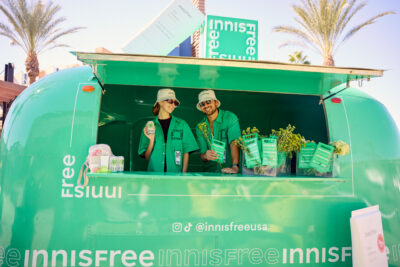 Two people in green shirts and beige hats hold products inside a green Innisfree trailer.