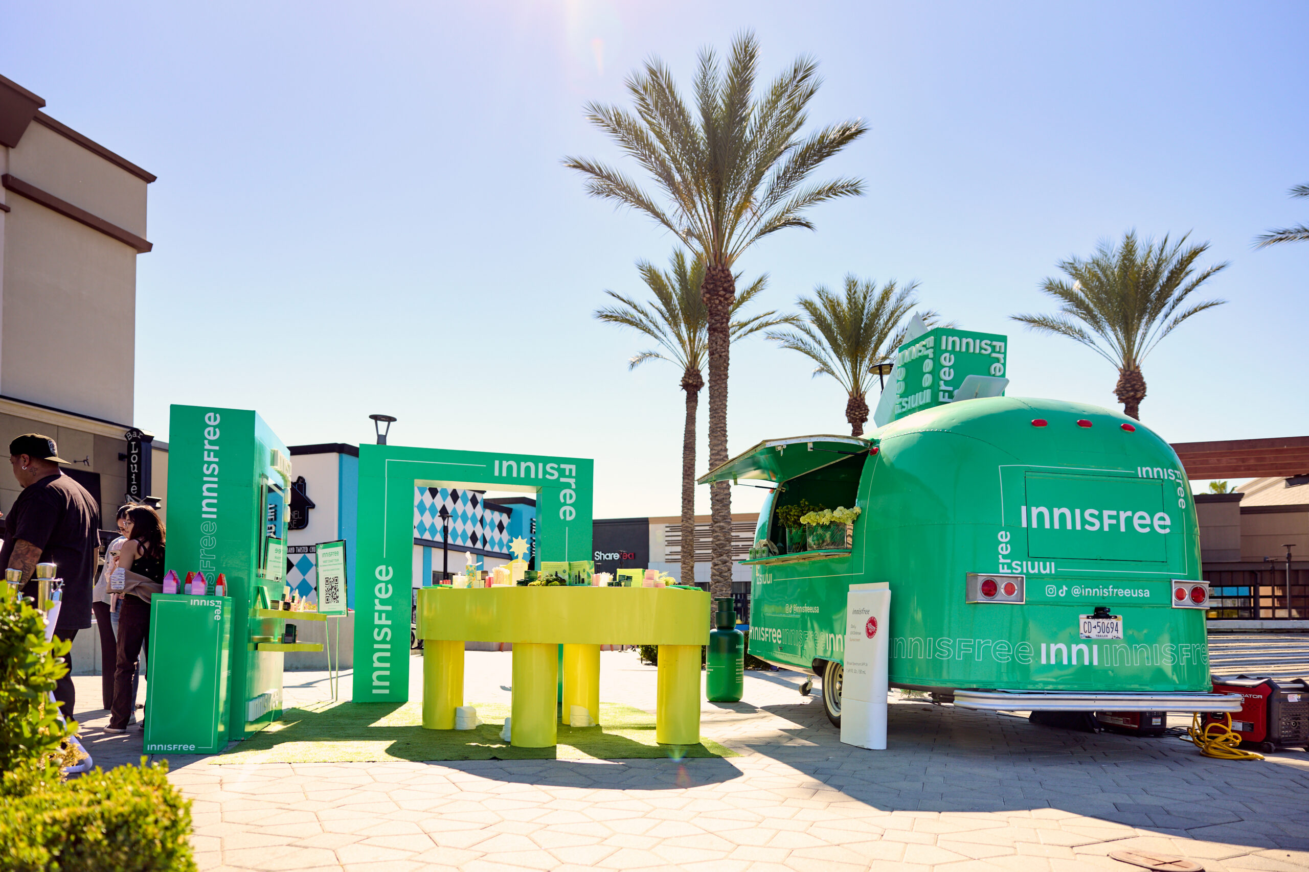 Innisfree pop-up with products, trailer, and tables outdoors by palm trees with several people.