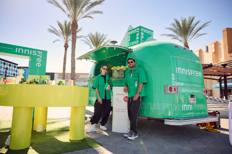 Two people in green shirts smile by a green Innisfree trailer and yellow product table outdoors.