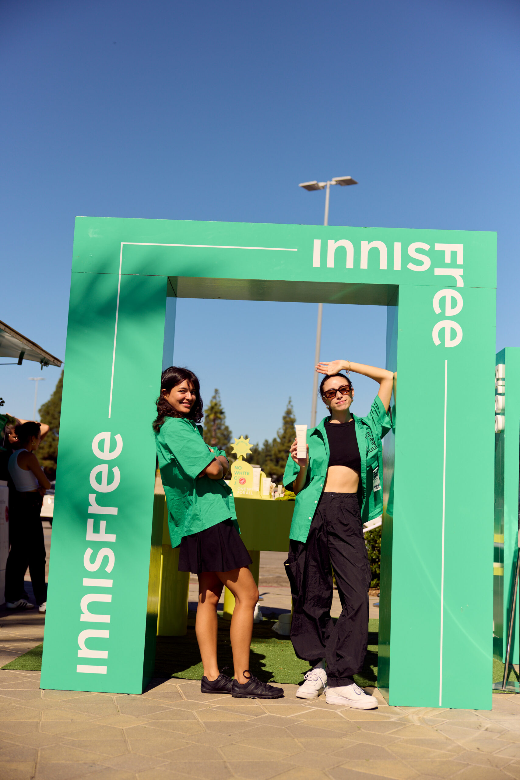 Two women in green shirts pose under a green Innisfree frame; one holds up a bottle, sky is clear.