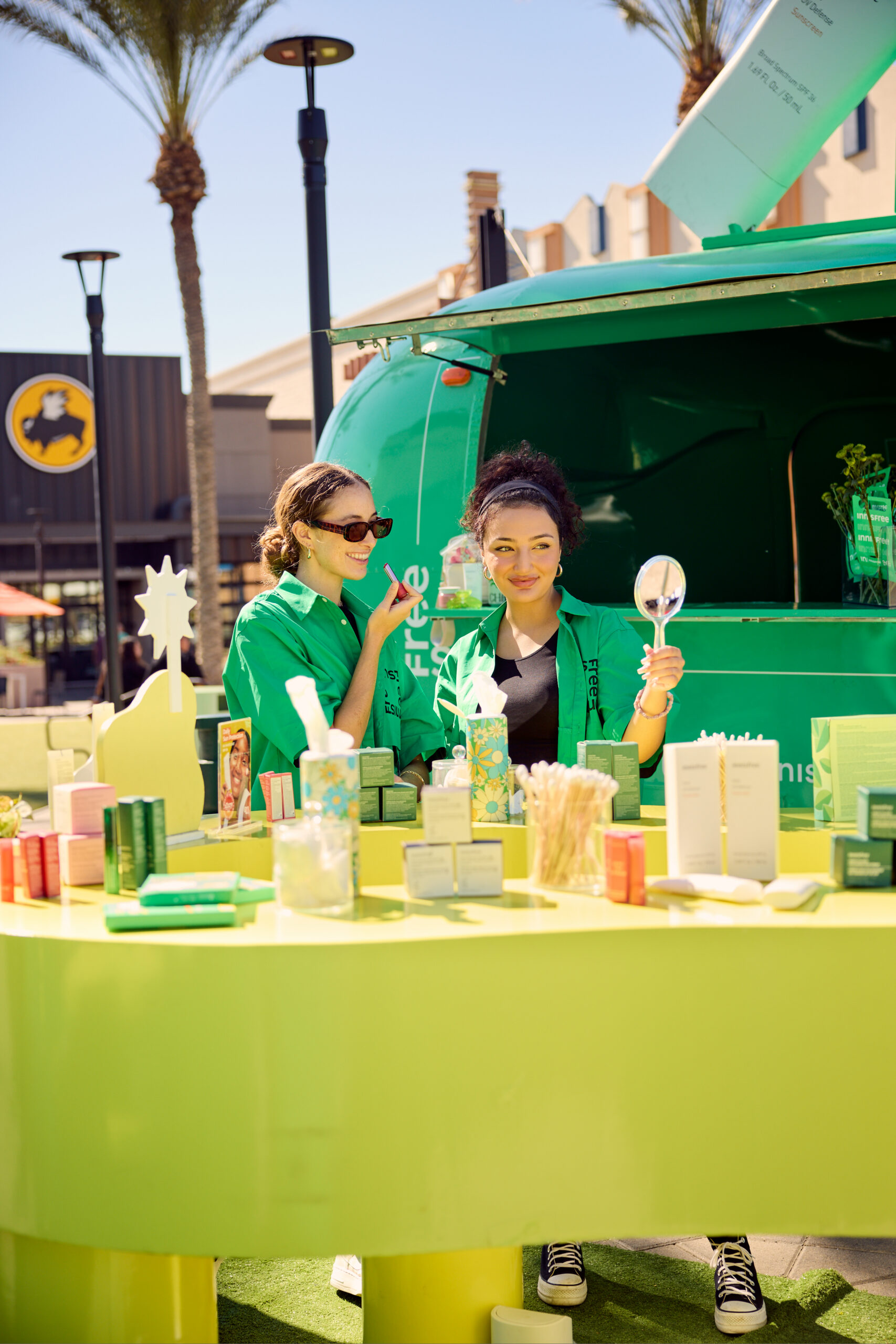 Two women in green shirts stand behind a table with skincare products; one smiles holding a mirror.