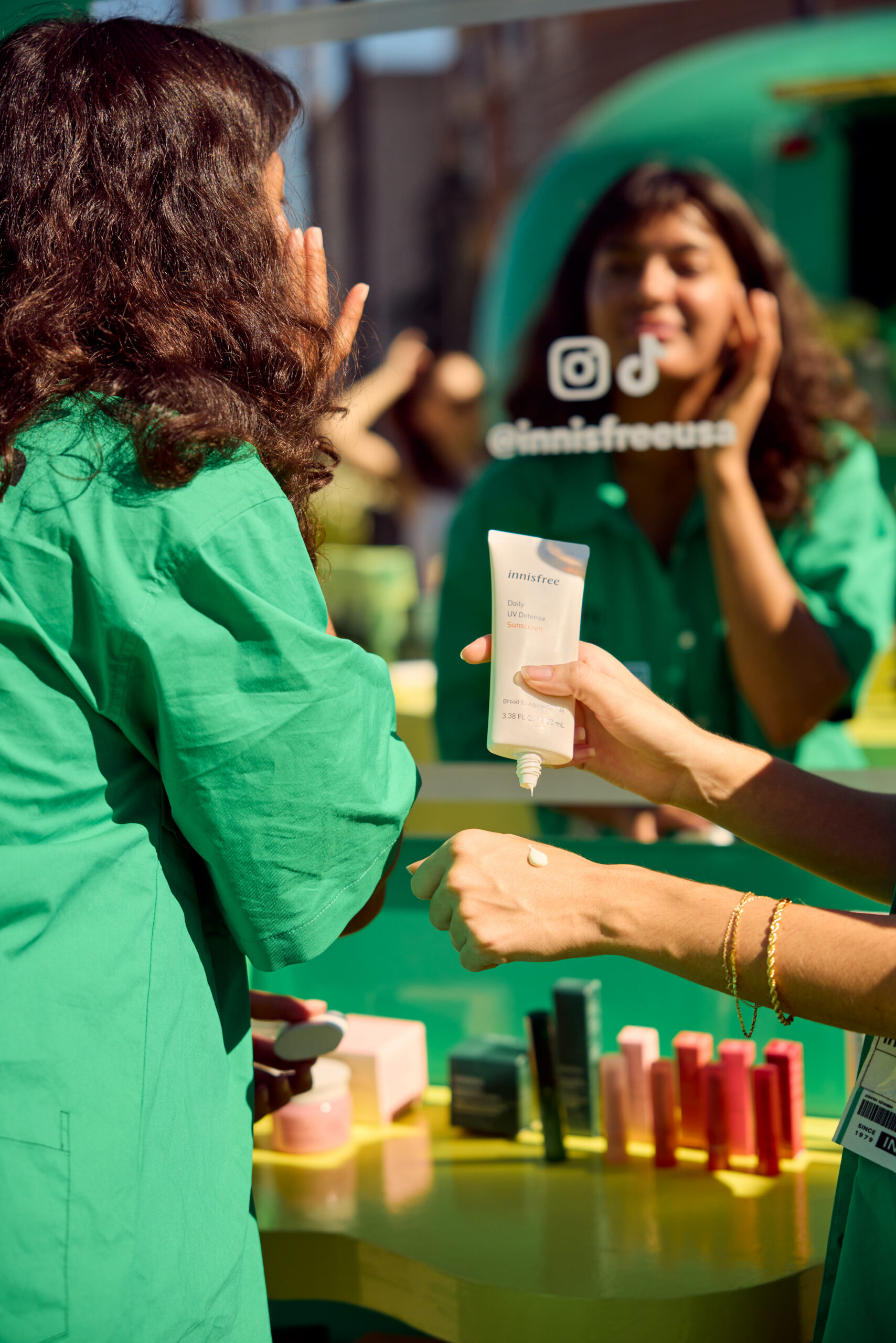 Woman in green shirt applies skincare cream by a mirror, with cosmetics on the counter.