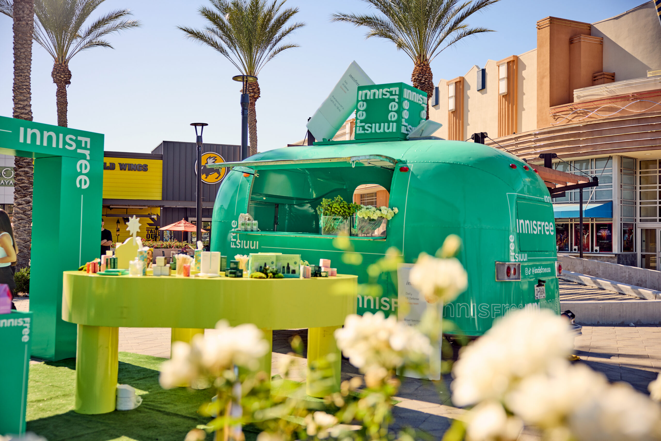 Green Innisfree pop-up trailer displays skincare on a round table, with palm trees and shops nearby.