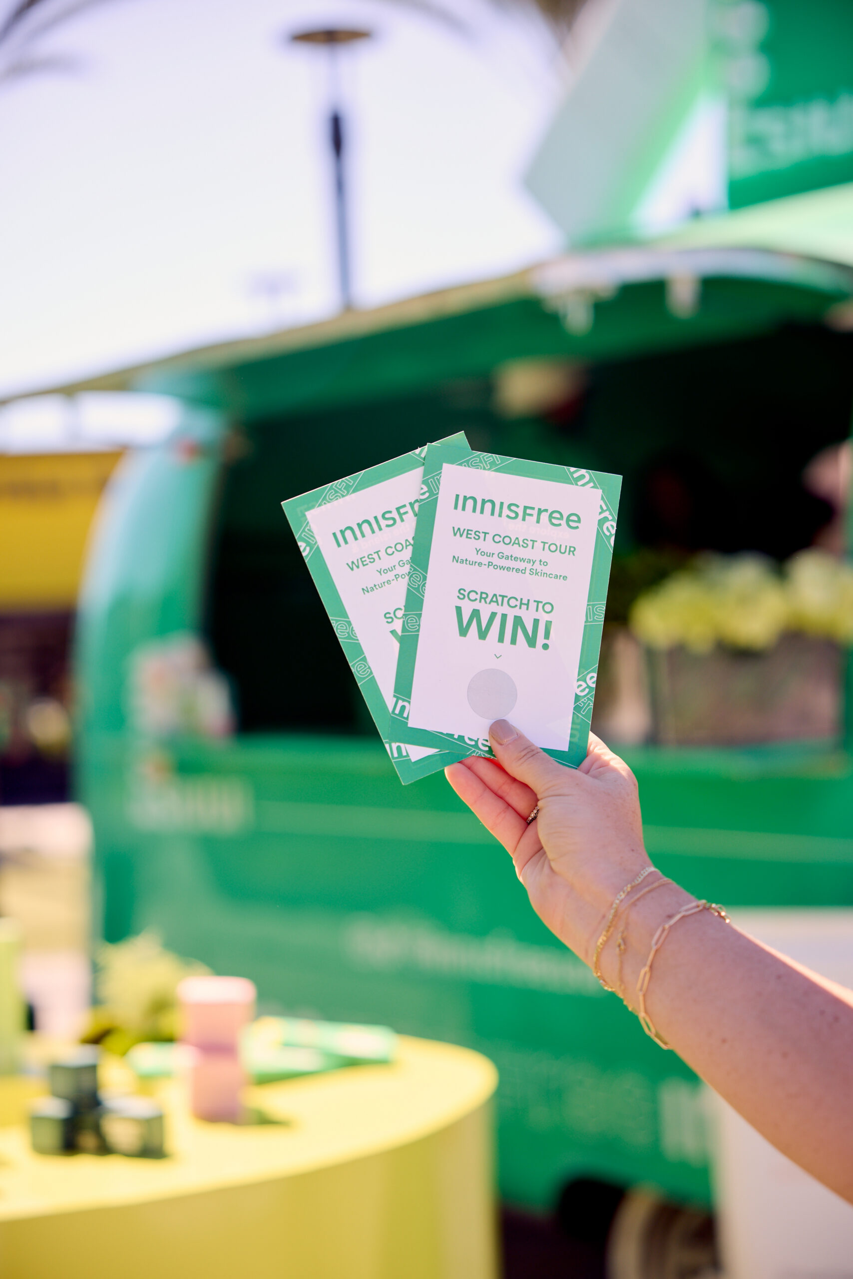 Hand holding two green Innisfree West Coast Tour scratch cards near a food truck and tables.