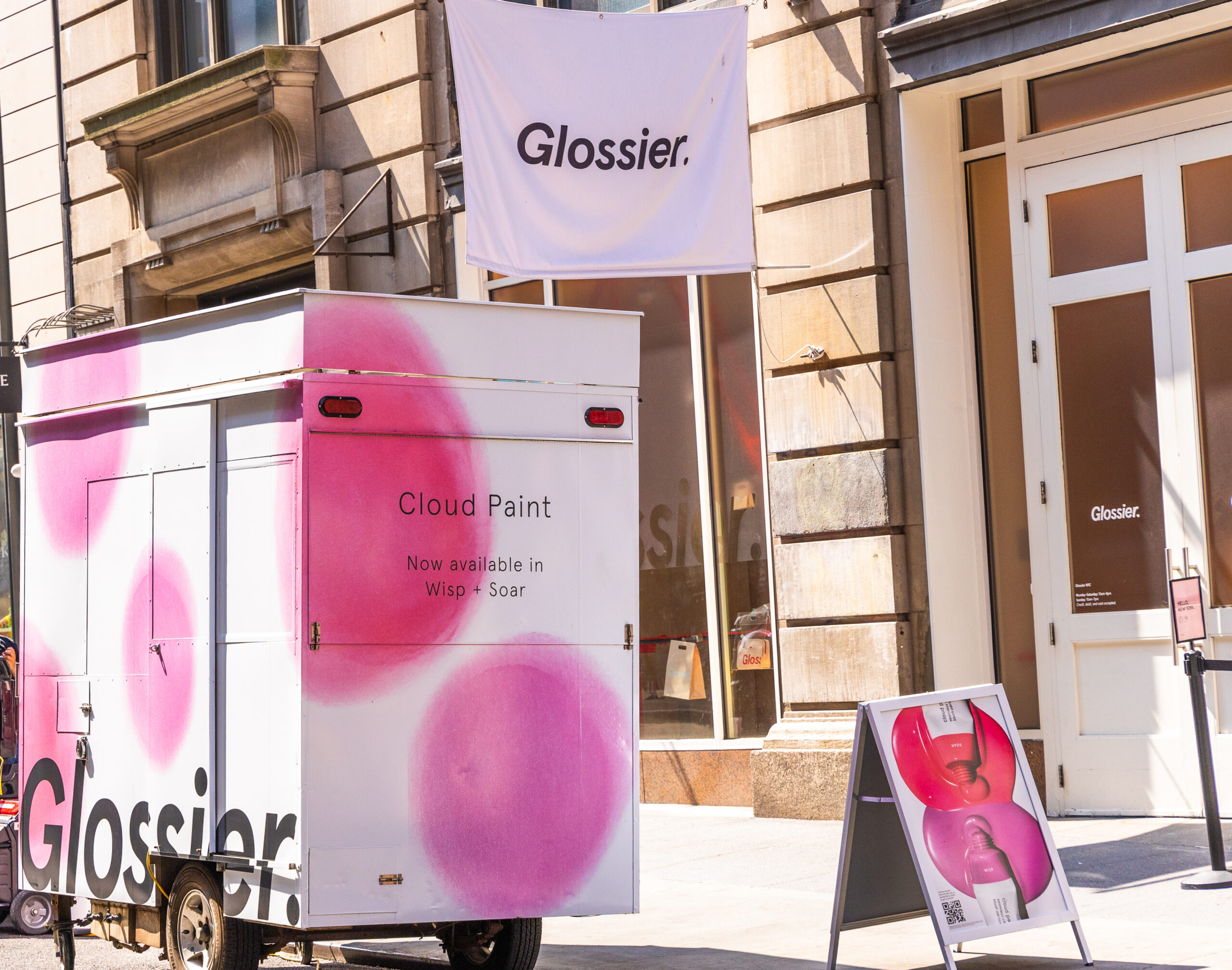 Glossier Cloud Paint pop-up with white and pink trailer, sign, and banner outside a building.