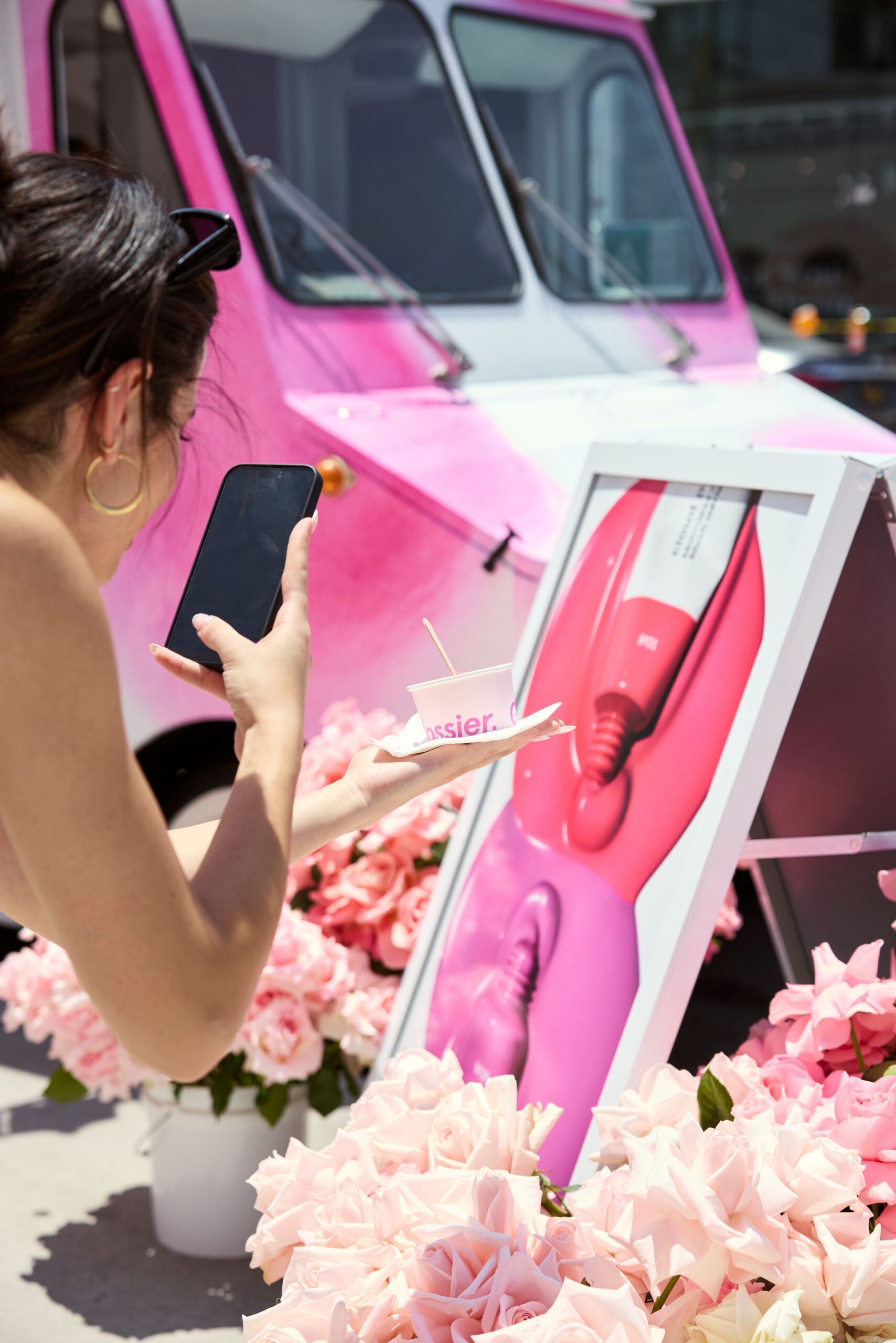 Woman takes a phone photo of a pink dessert cup at a cloud paint pop-up with an ice cream truck.