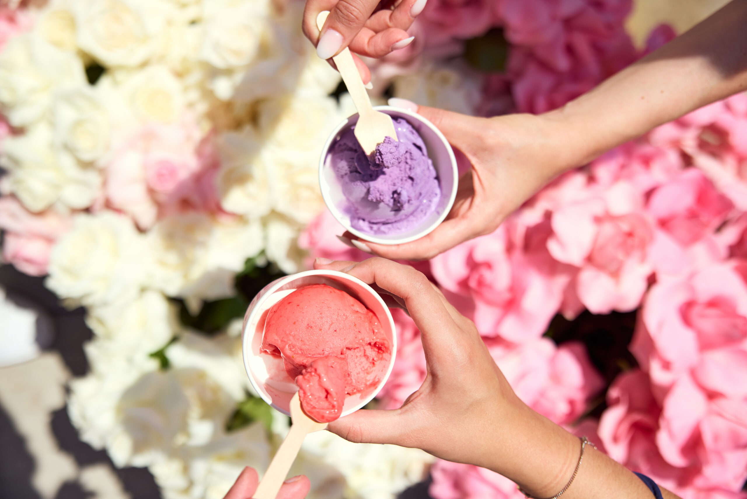 Two people hold purple and pink ice cream cups with wooden spoons, flowers in the background.