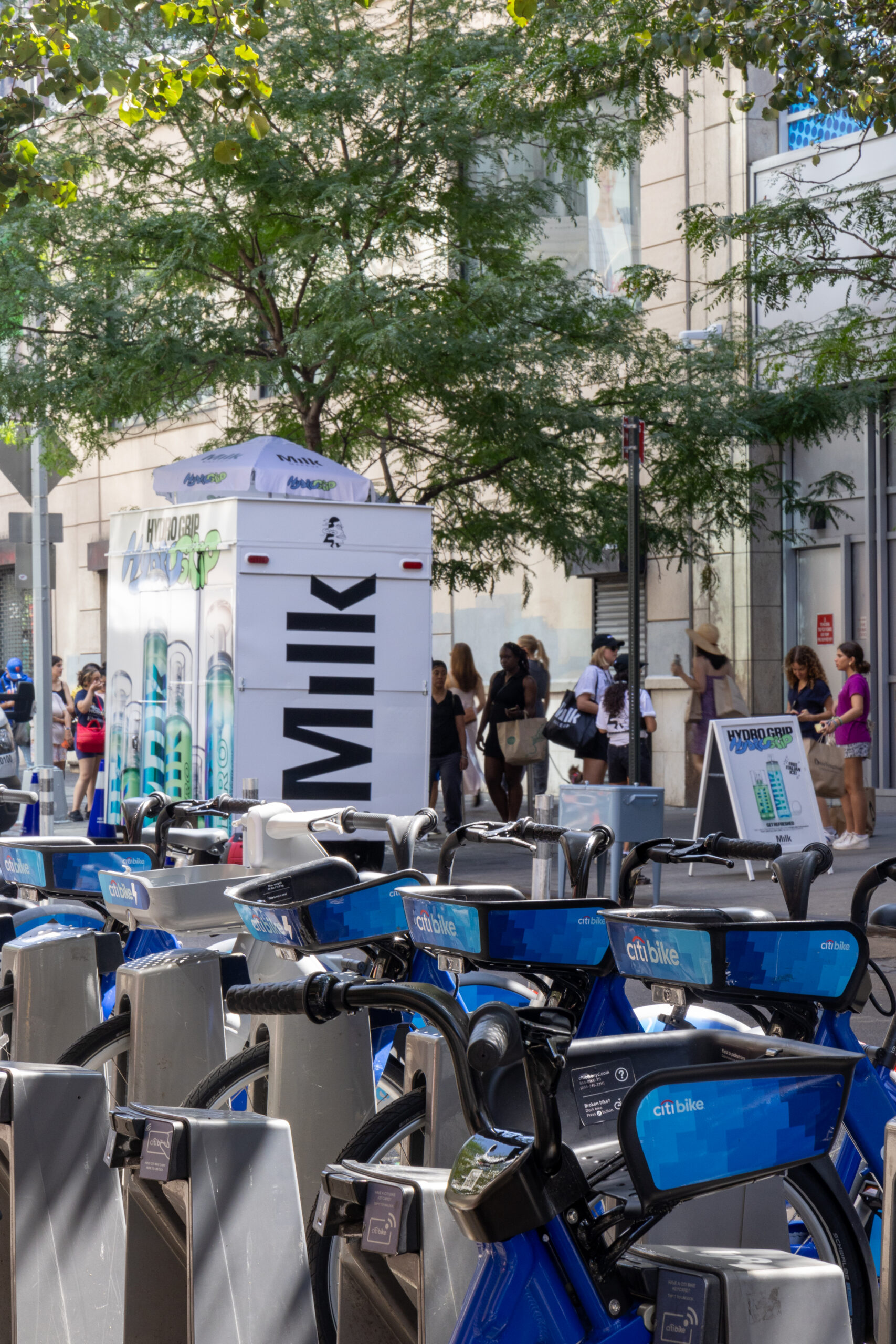 Blue Citi Bikes parked at a bike station, people in line near a white Milk truck on the sidewalk.