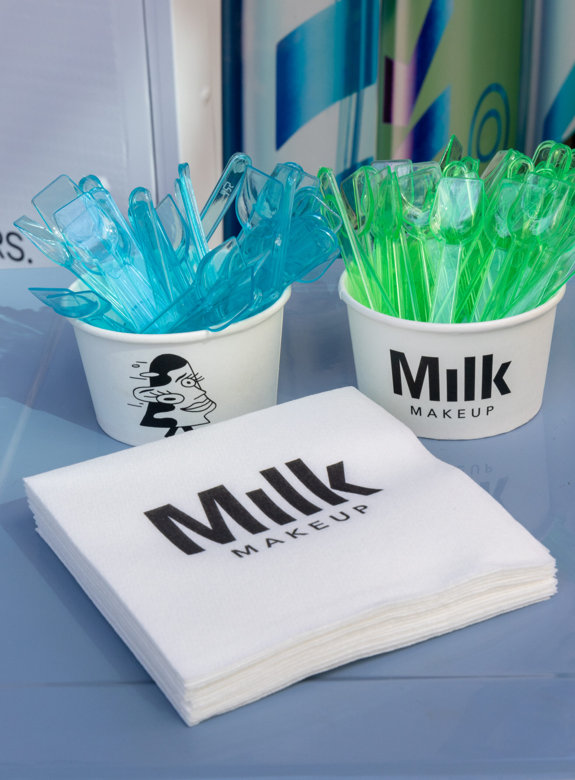 Two Milk Makeup cups with blue and green spatulas, and branded napkins on a blue surface.