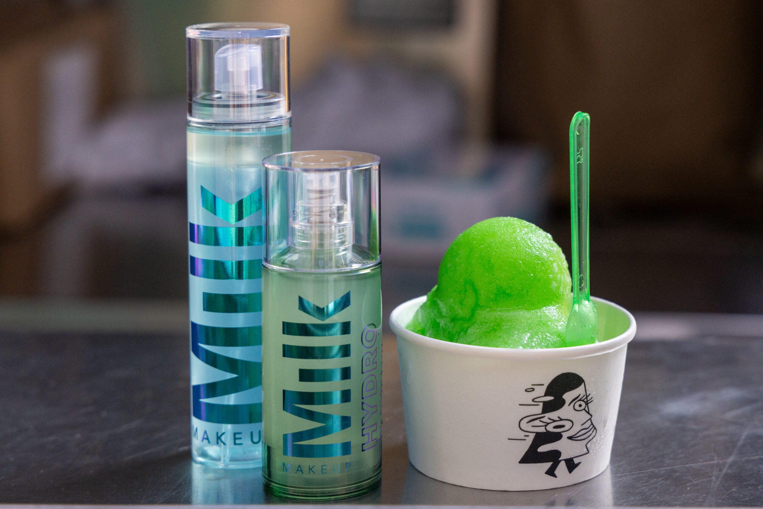 Two MILK Makeup Hydro bottles beside a cup of green sorbet with a clear spoon and cartoon design.