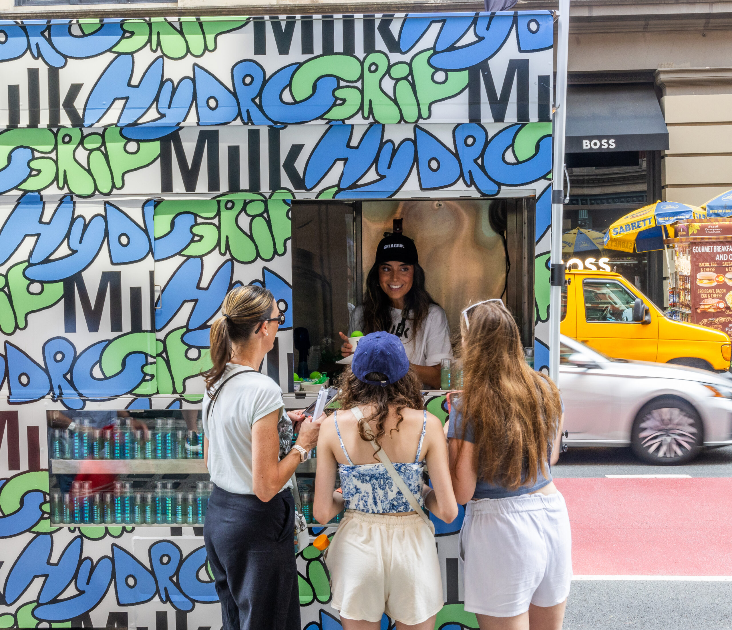 Three women talk to a smiling worker at a food truck with "Hydro" graffiti; city traffic behind.