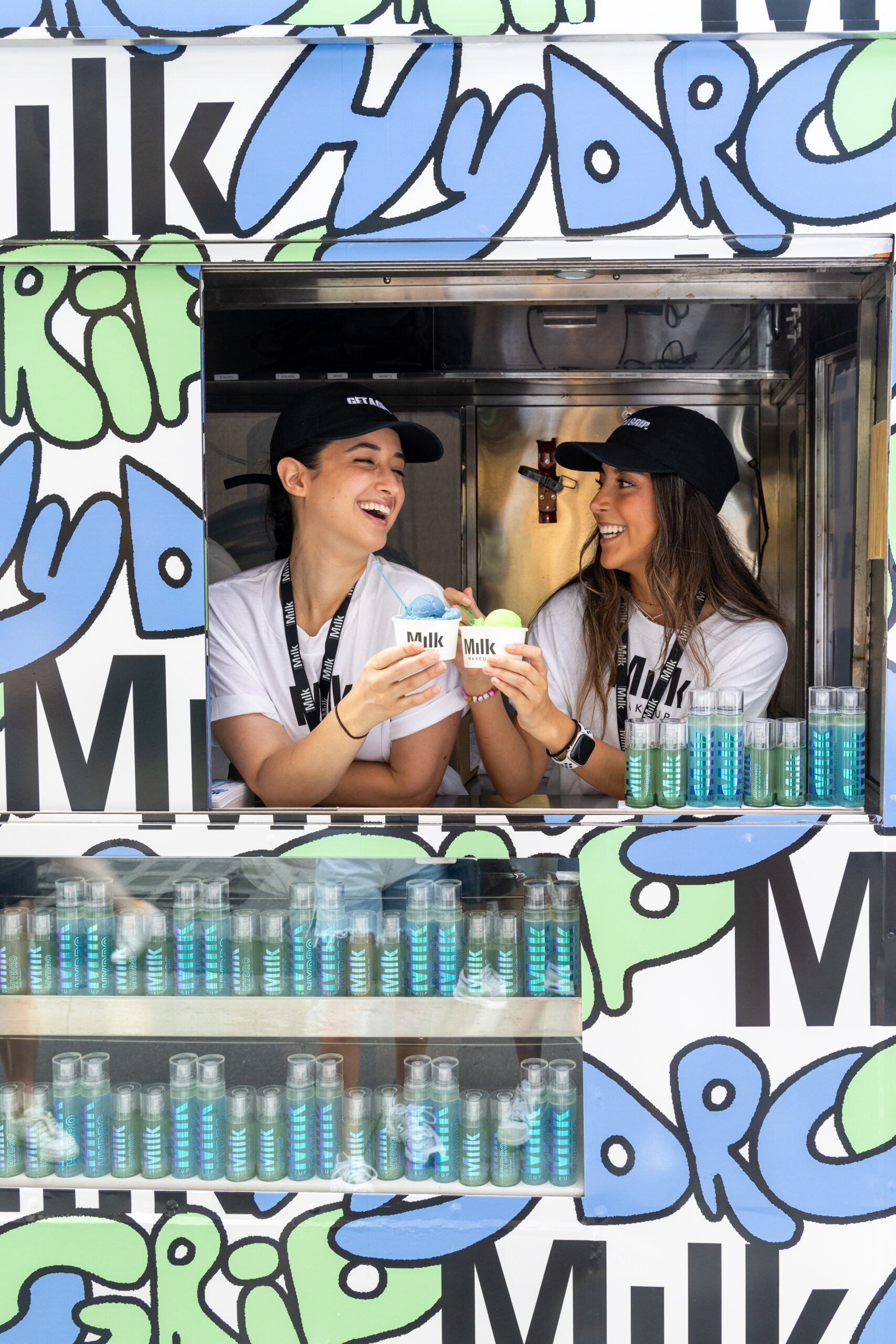 Two women in black hats toast with milk cartons inside a food truck window lined with bottles.