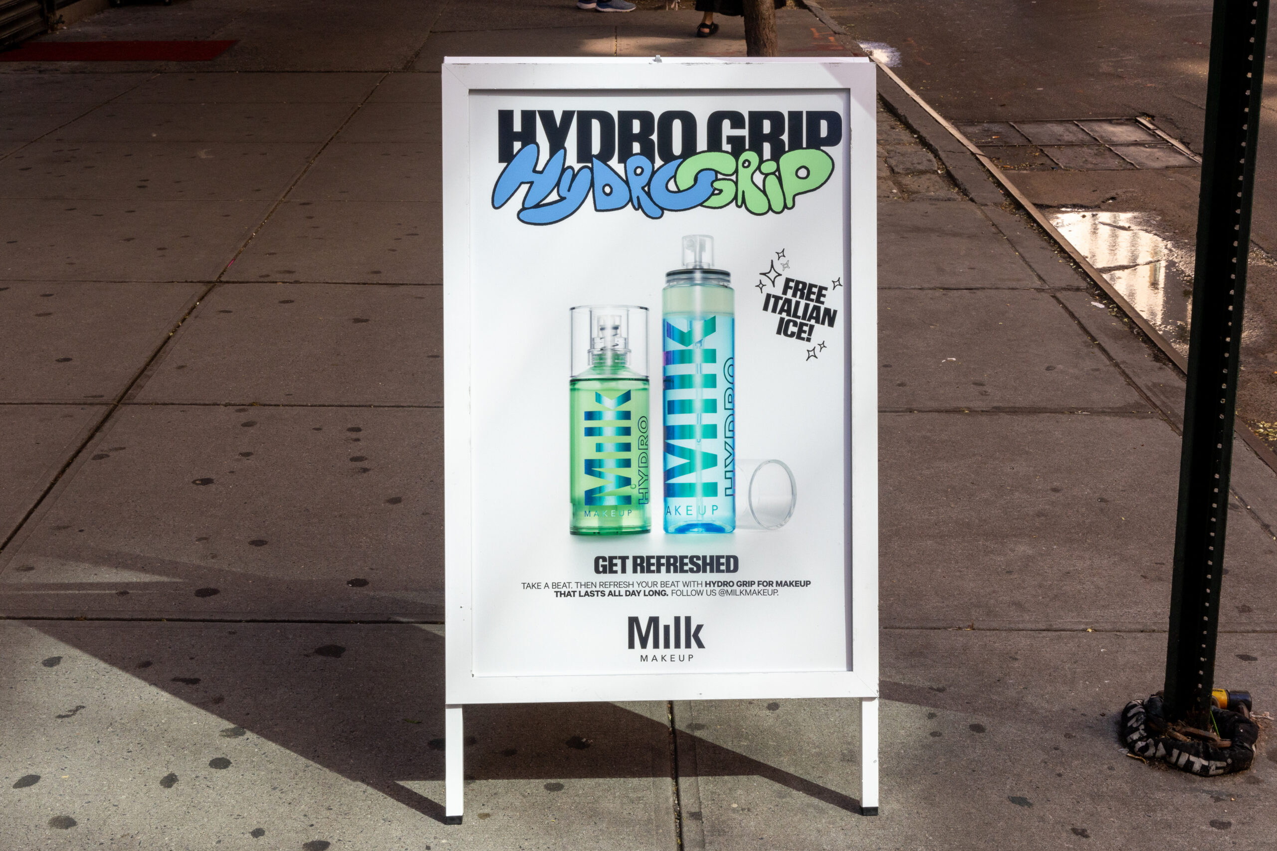 Sidewalk sign with two Milk Makeup Hydro Grip bottles and "free Italian ice" text by a streetlamp.