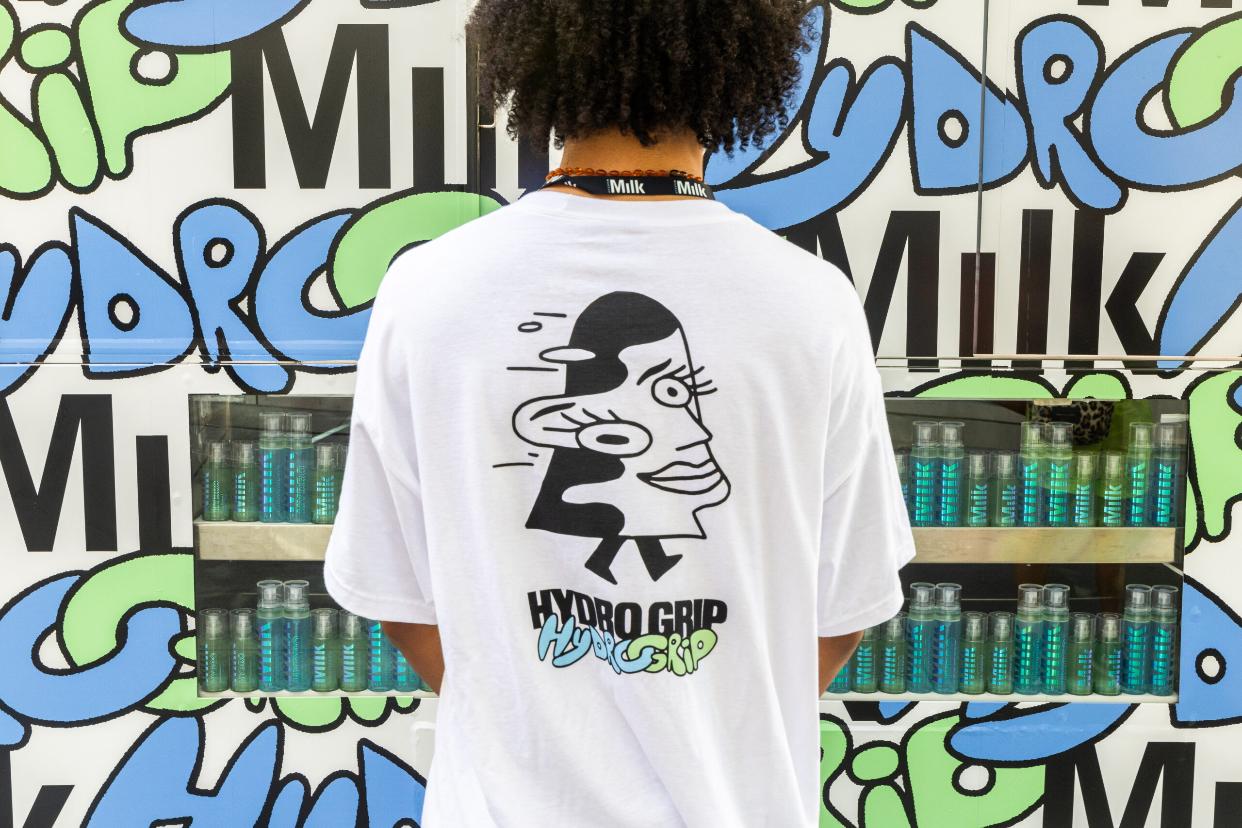 Person with curly hair faces a graffiti mural; wears a white HYDRO GRIP shirt. Bottles on shelves.