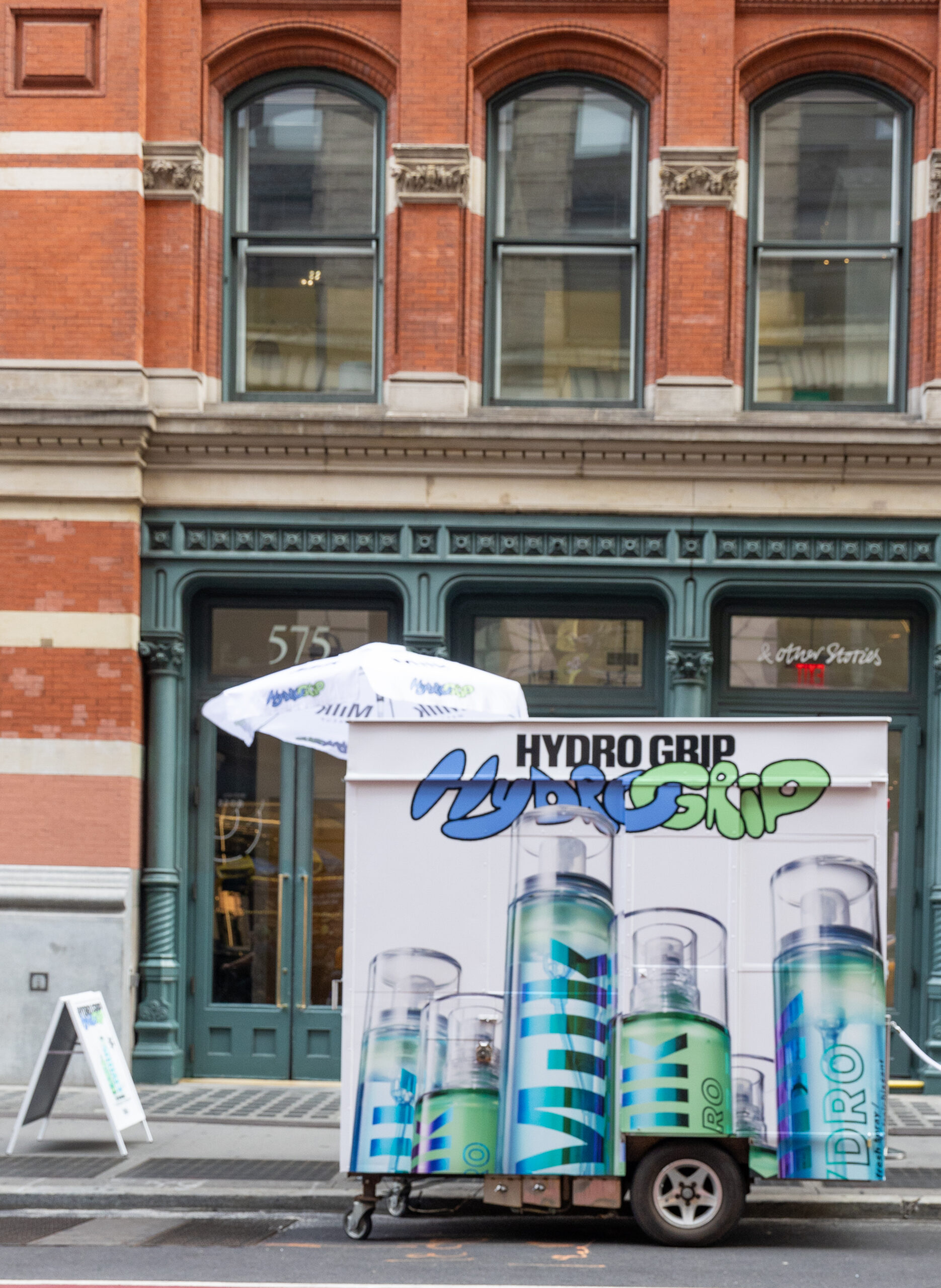 A street cart with Hydro Grip bottle images and a branded umbrella is by a brick building.
