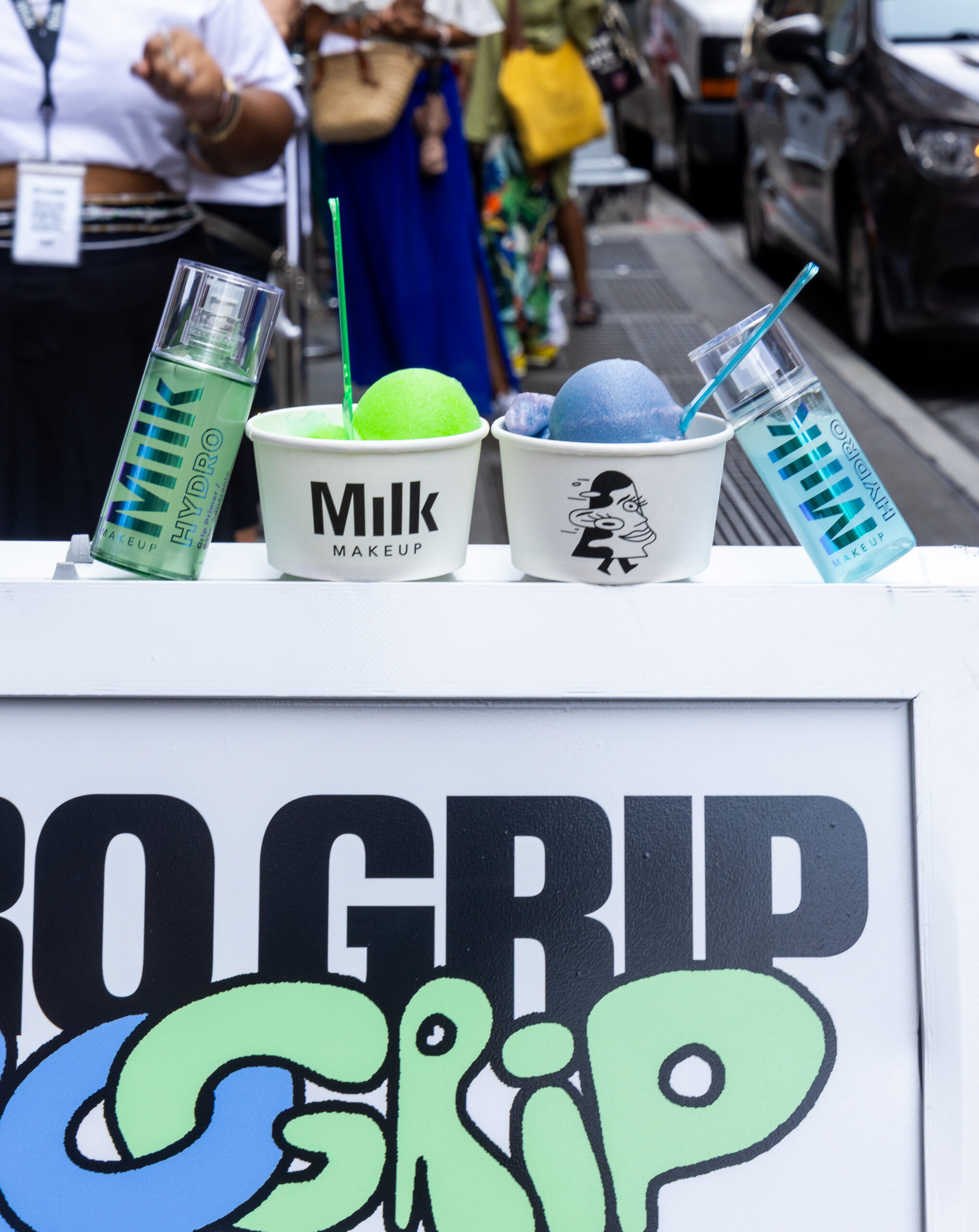 Two Milk Makeup Hydro Grip bottles and two cups of shaved ice on a white outdoor sign.