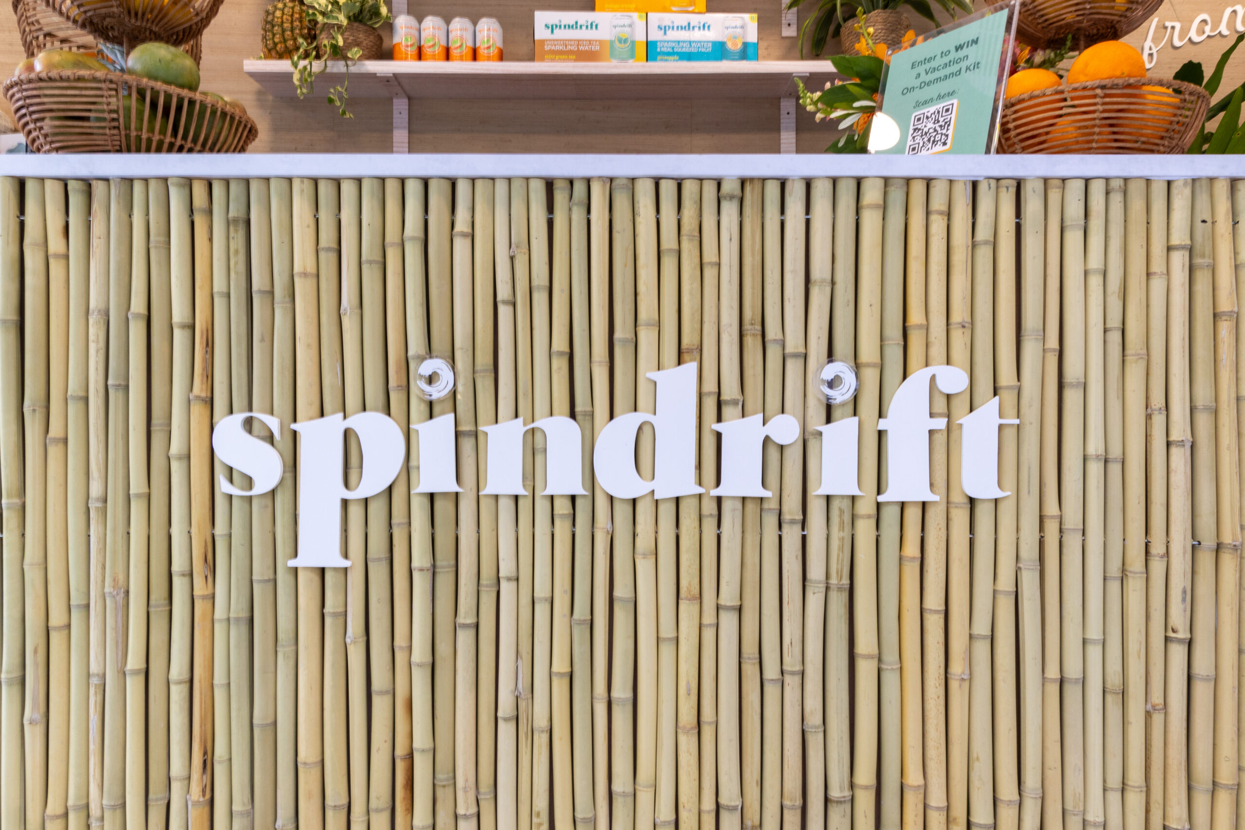 Bamboo counter labeled spindrift displays fruit baskets, drink cans, and a QR code sign in back.