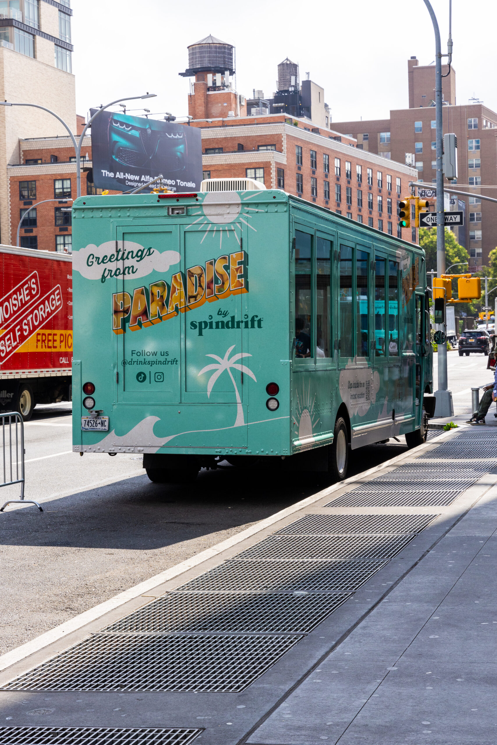 Turquoise Spindrift pop-up bus with palm tree graphics parked on a city street by buildings.