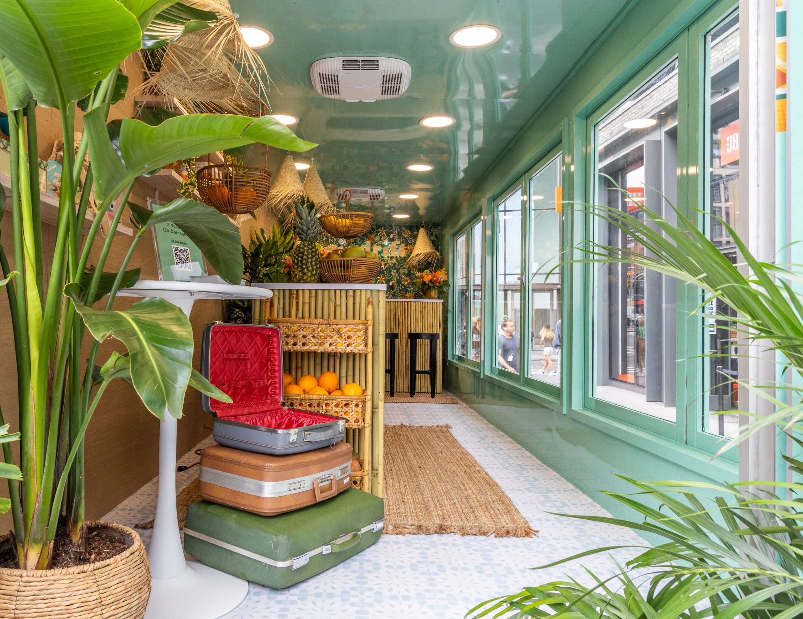 Tropical pop-up room with large windows, green plants, wicker decor, oranges, and a woven rug.