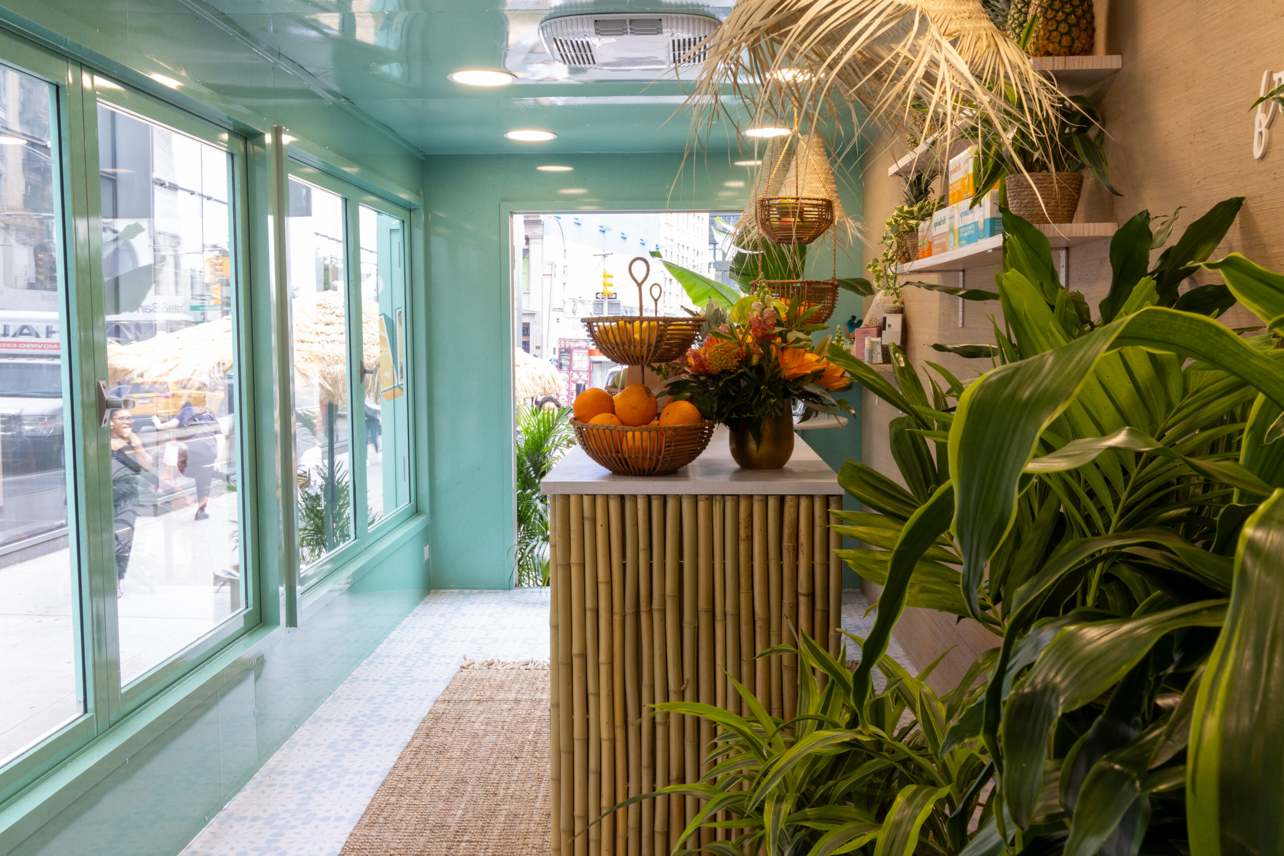 Tropical pop-up room with large windows, bamboo counter with fruit and flowers, plants, and shelves.