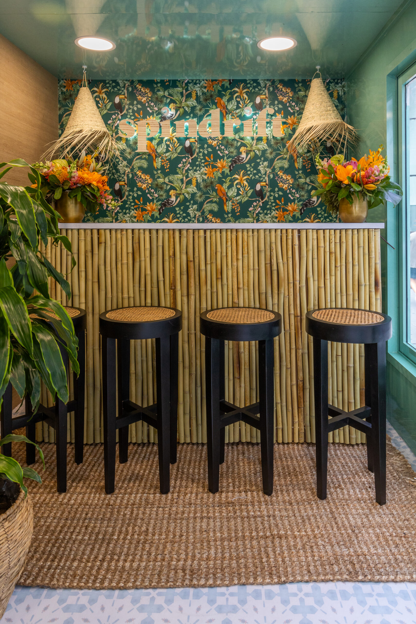 Tropical pop-up bar with bamboo counter, four black stools, floral decor, and a plant on a rug.