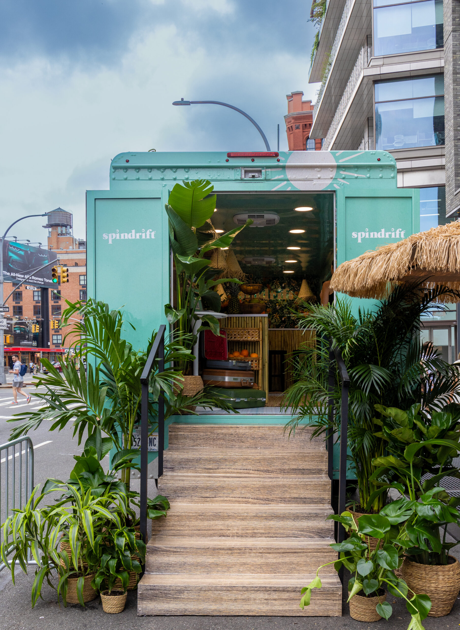 Green Spindrift pop-up truck with plants and wicker furniture parked on a busy city street.