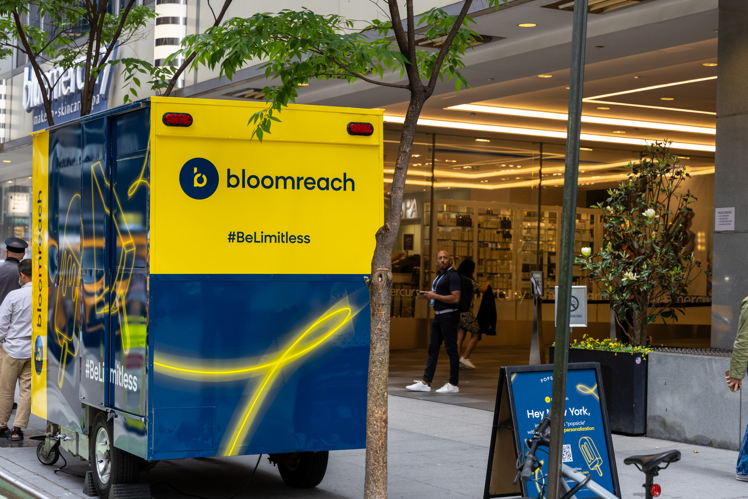 A blue and yellow Bloomreach popsicle cart is parked on a city sidewalk near a glass storefront.