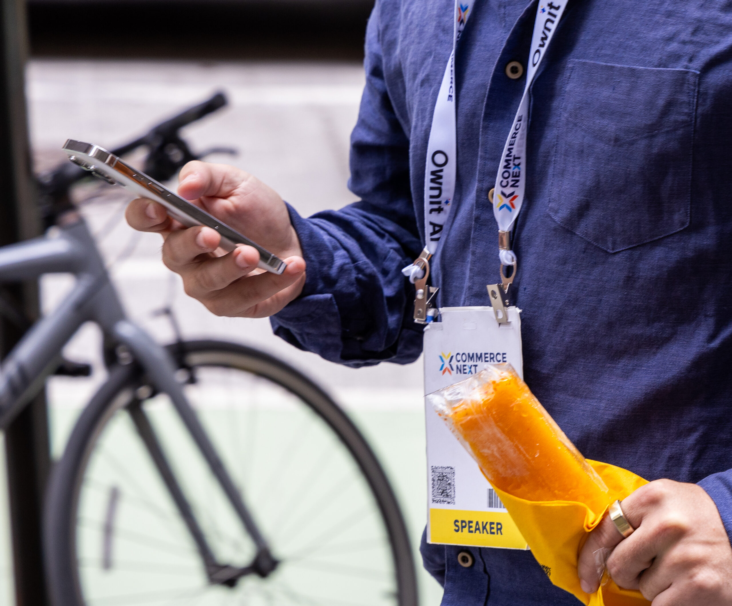 Person in blue shirt holds phone and orange ice pop, wearing SPEAKER badge; bicycle in background.